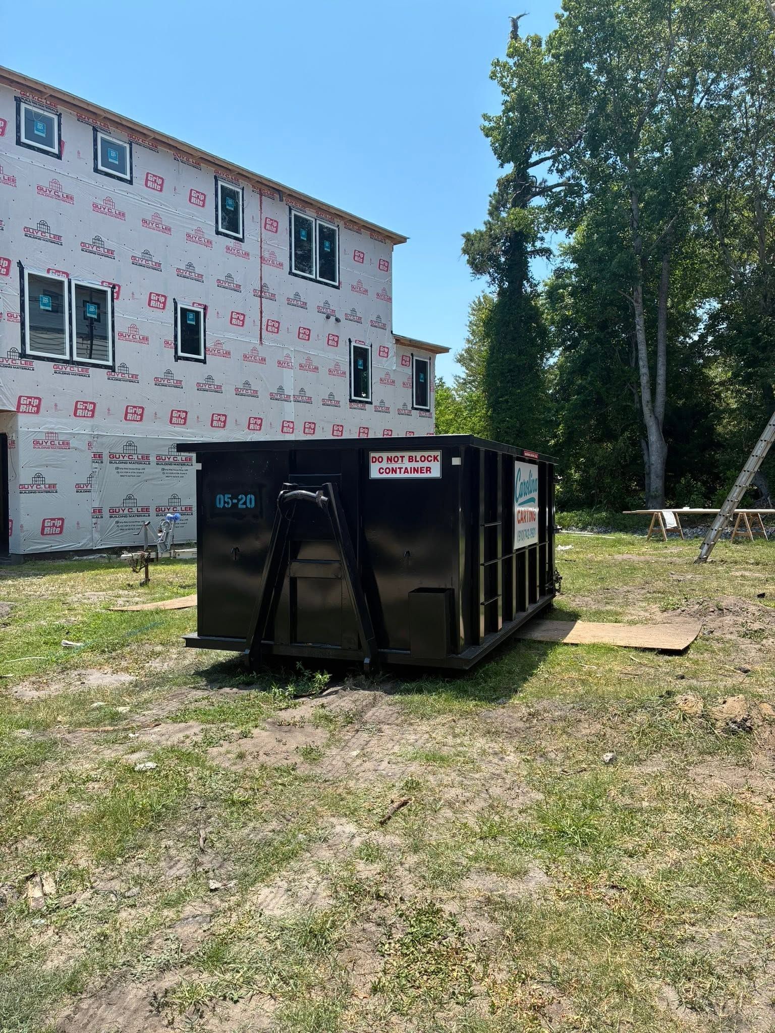 Black dumpster on grass beside a partially finished white house and tall trees