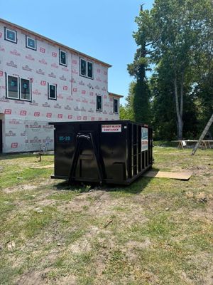 Black dumpster on grass beside a partially finished white house and tall trees