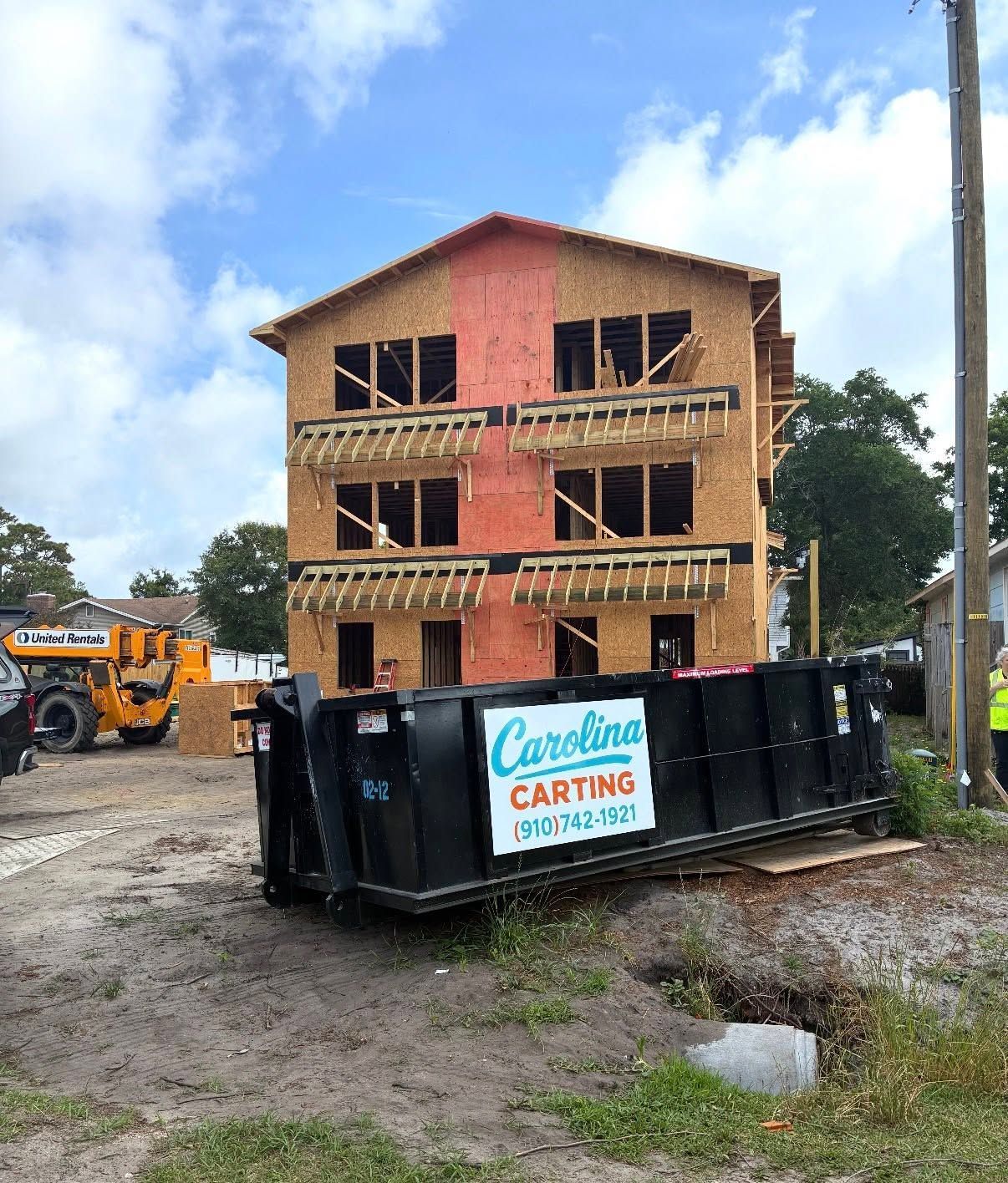 Construction site with unfinished brick building, overturned black dumpster, and excavator on muddy ground