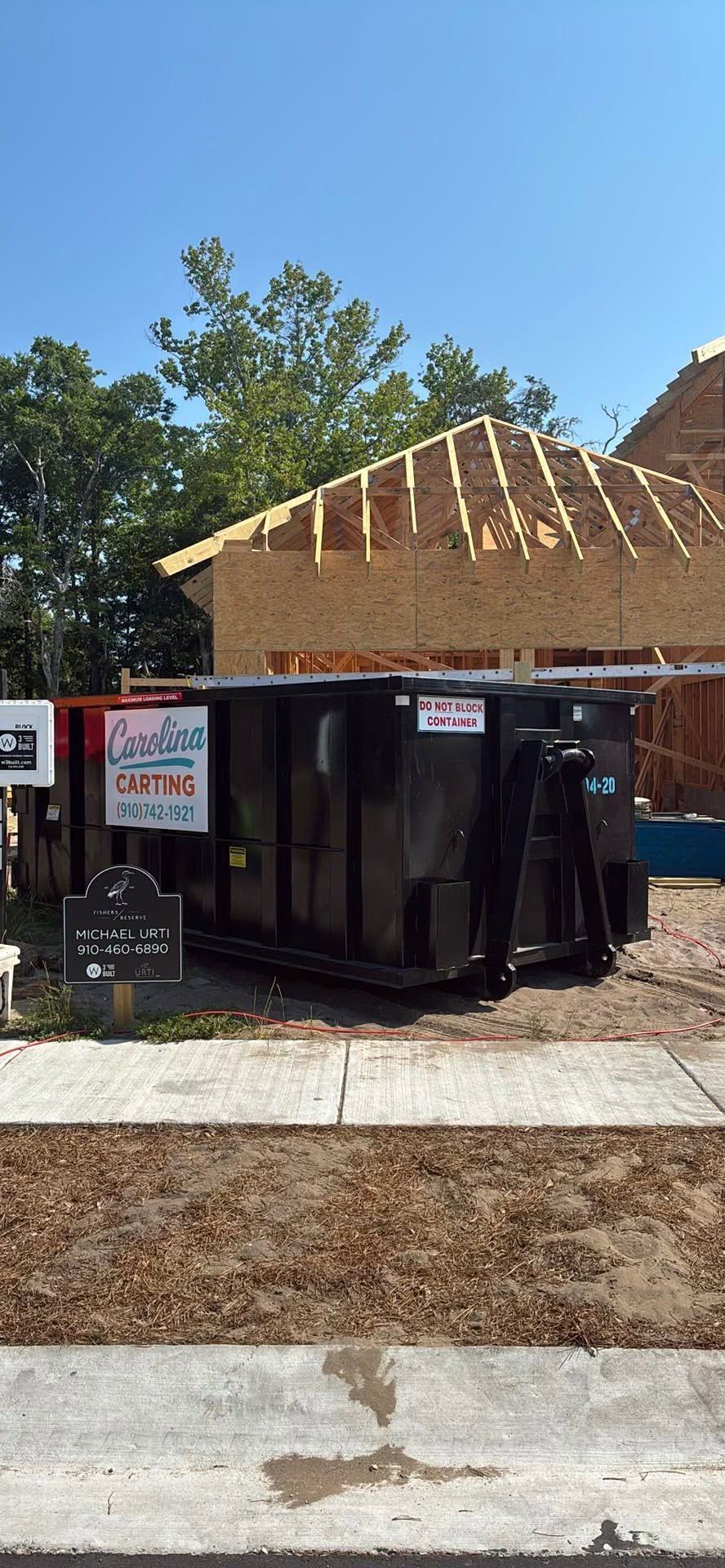 Construction dumpster outside a house under renovation, with exposed roof framing and a blue sky.