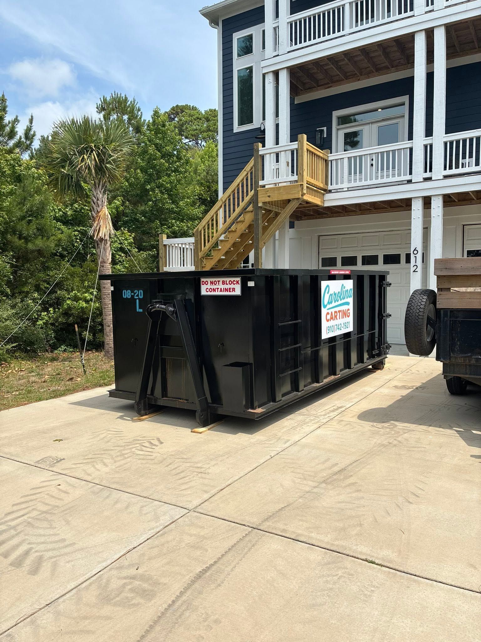 Black construction dumpster on a driveway beside a blue house with white railings and stairs