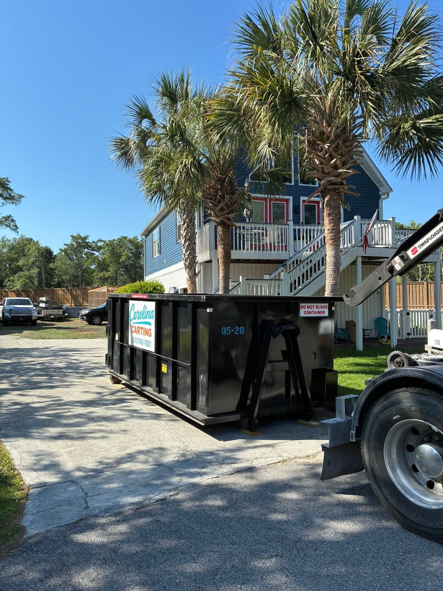 Black dumpster beside a beach house and palm trees on a sunny day
