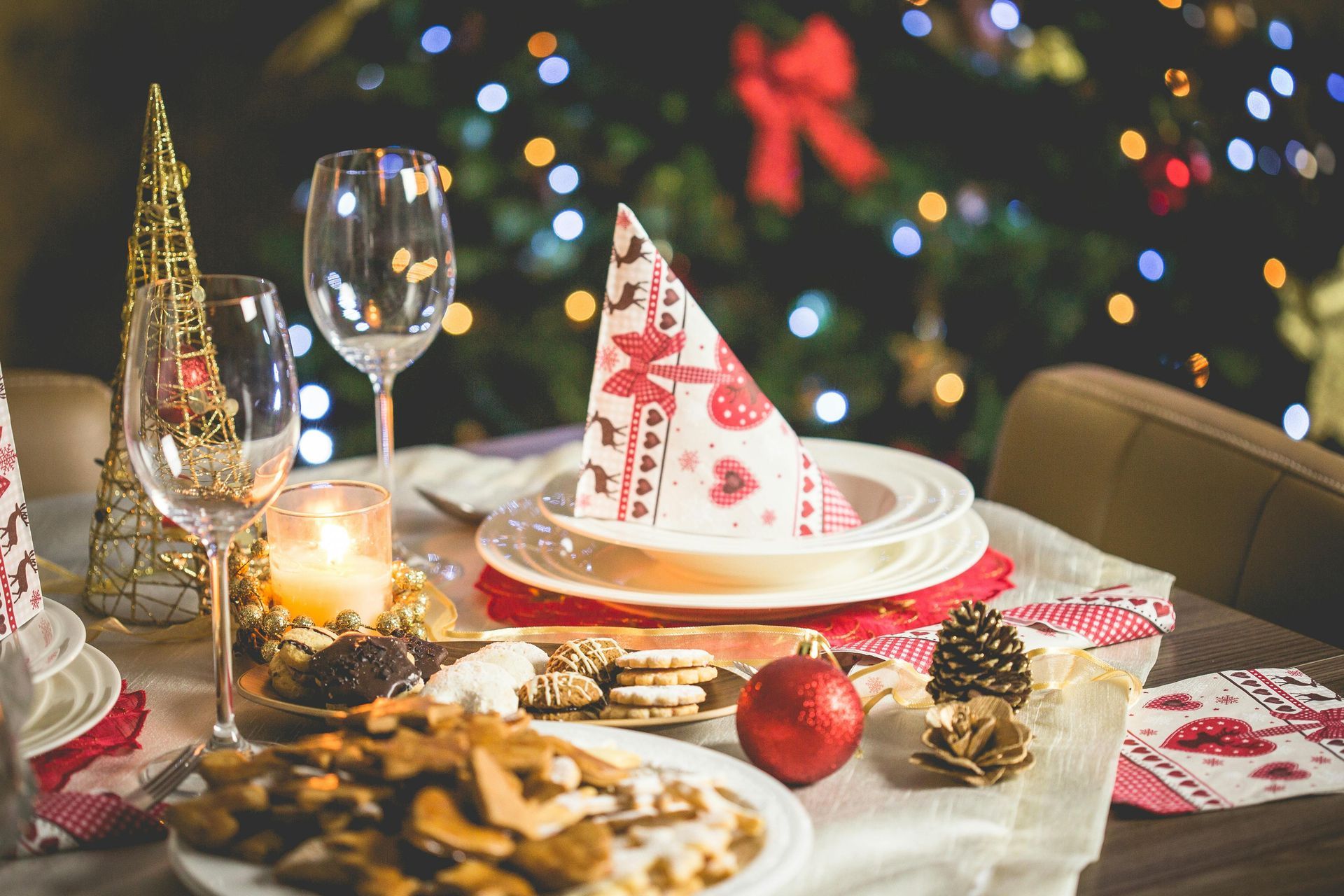 Christmas dinner table set with decorated plates, food, glasses, and tree in the background.