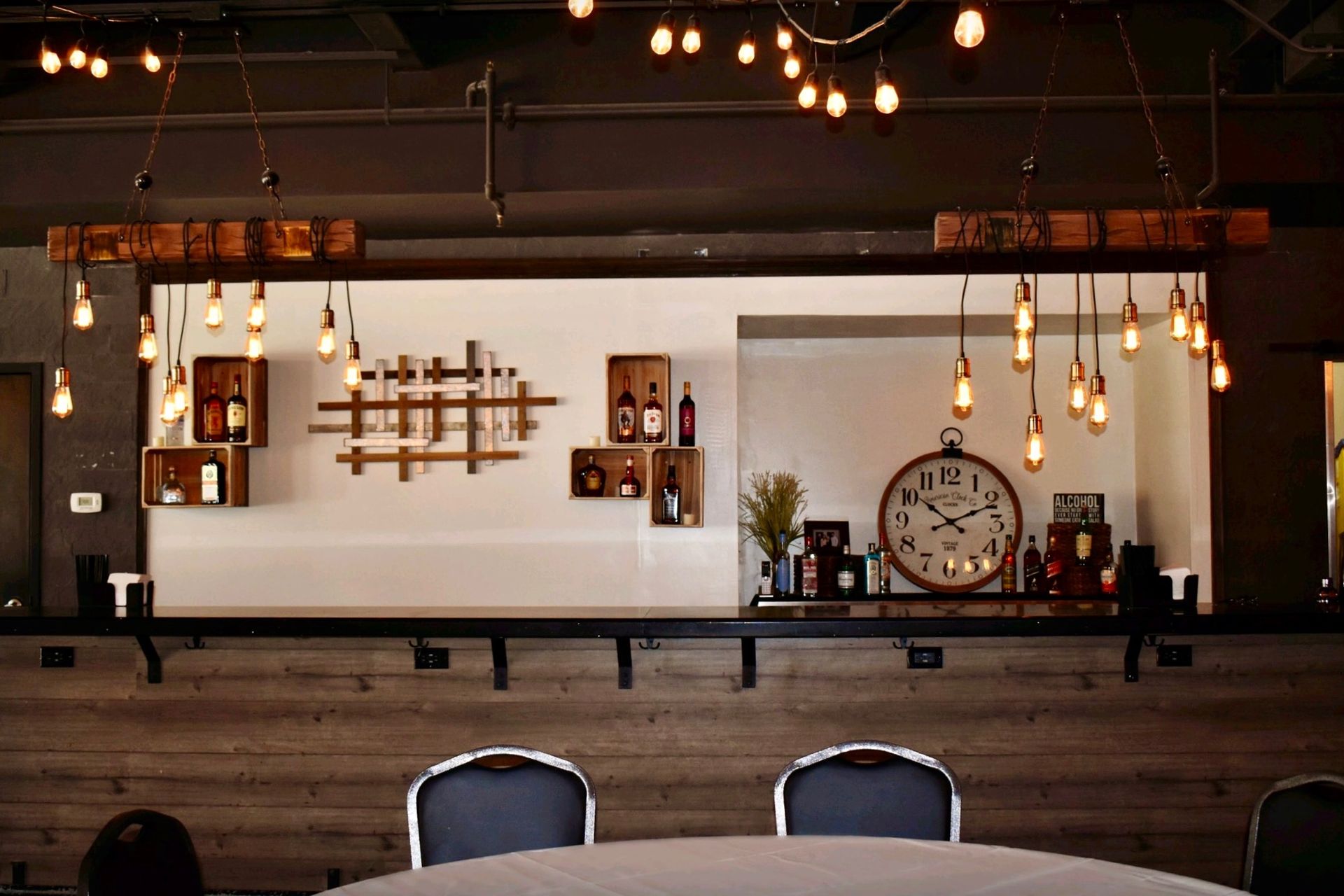 Bar interior with rustic decor: wooden bar, hanging lights, wine rack, and a large clock.