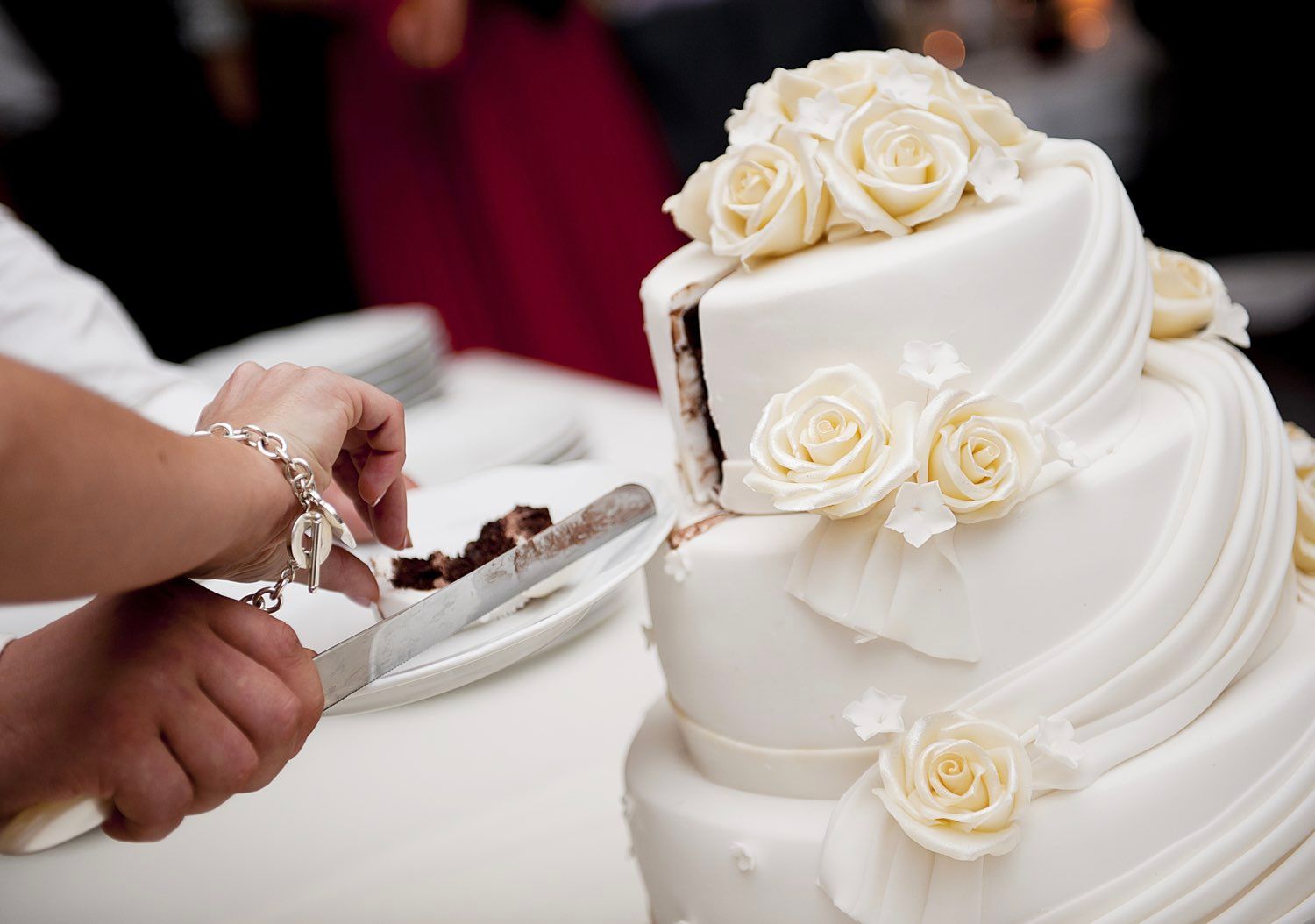 Person cutting into a white, tiered wedding cake decorated with roses.