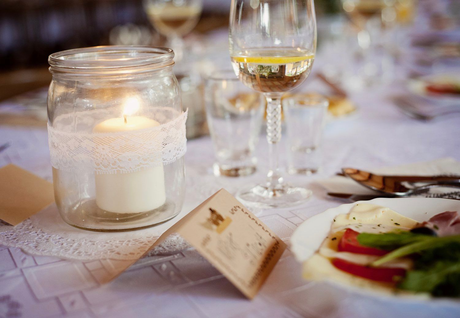 Wedding table setting with lit candle in jar, wine glasses, and plates of food.