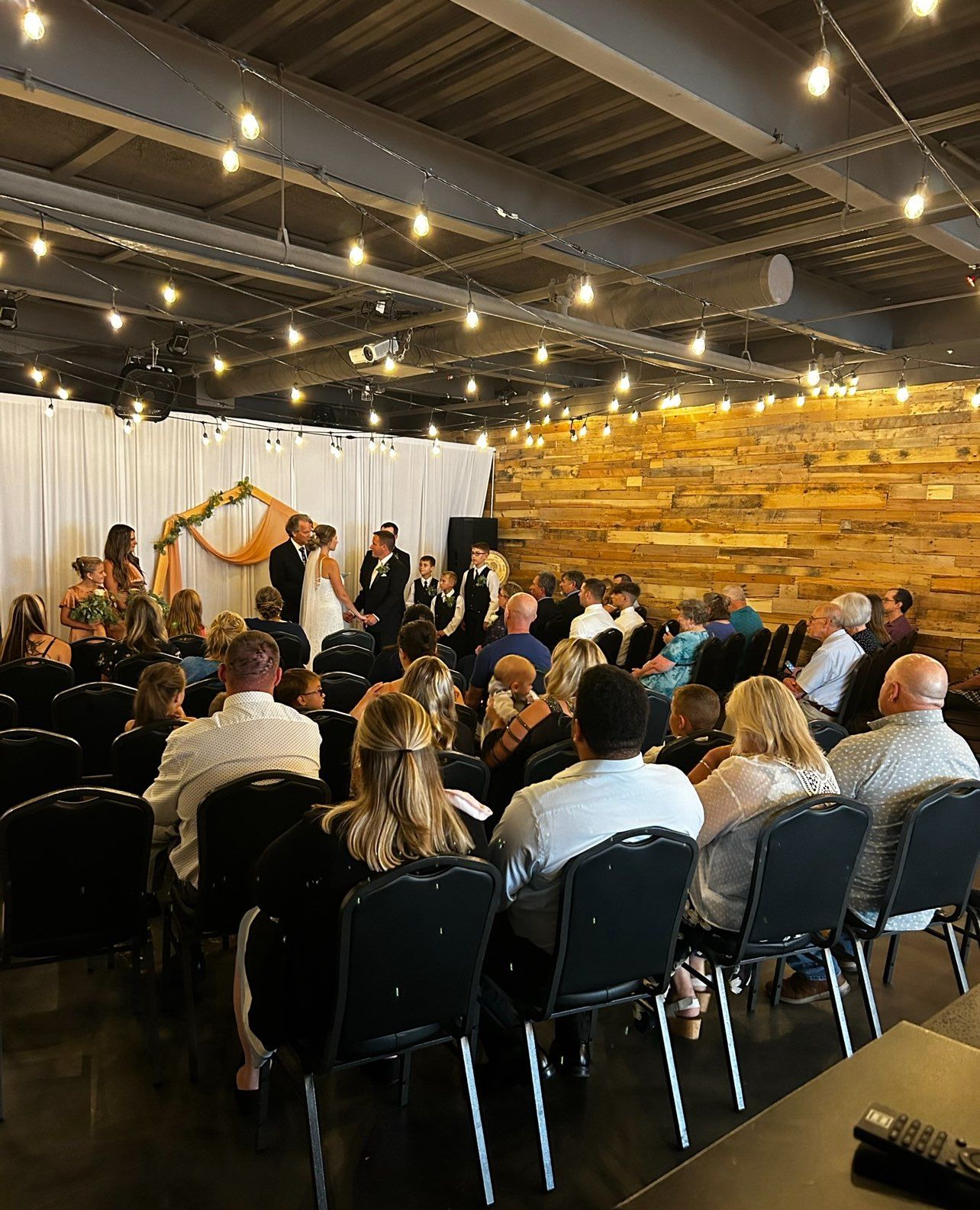 Wedding ceremony in an indoor space with guests seated, bride and groom at the altar, and a backdrop of lights and decor.