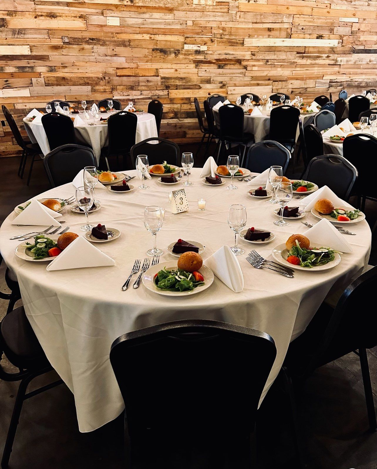 Round tables set for a formal event with white tablecloths, food, and black chairs in a room with a wood-paneled wall.