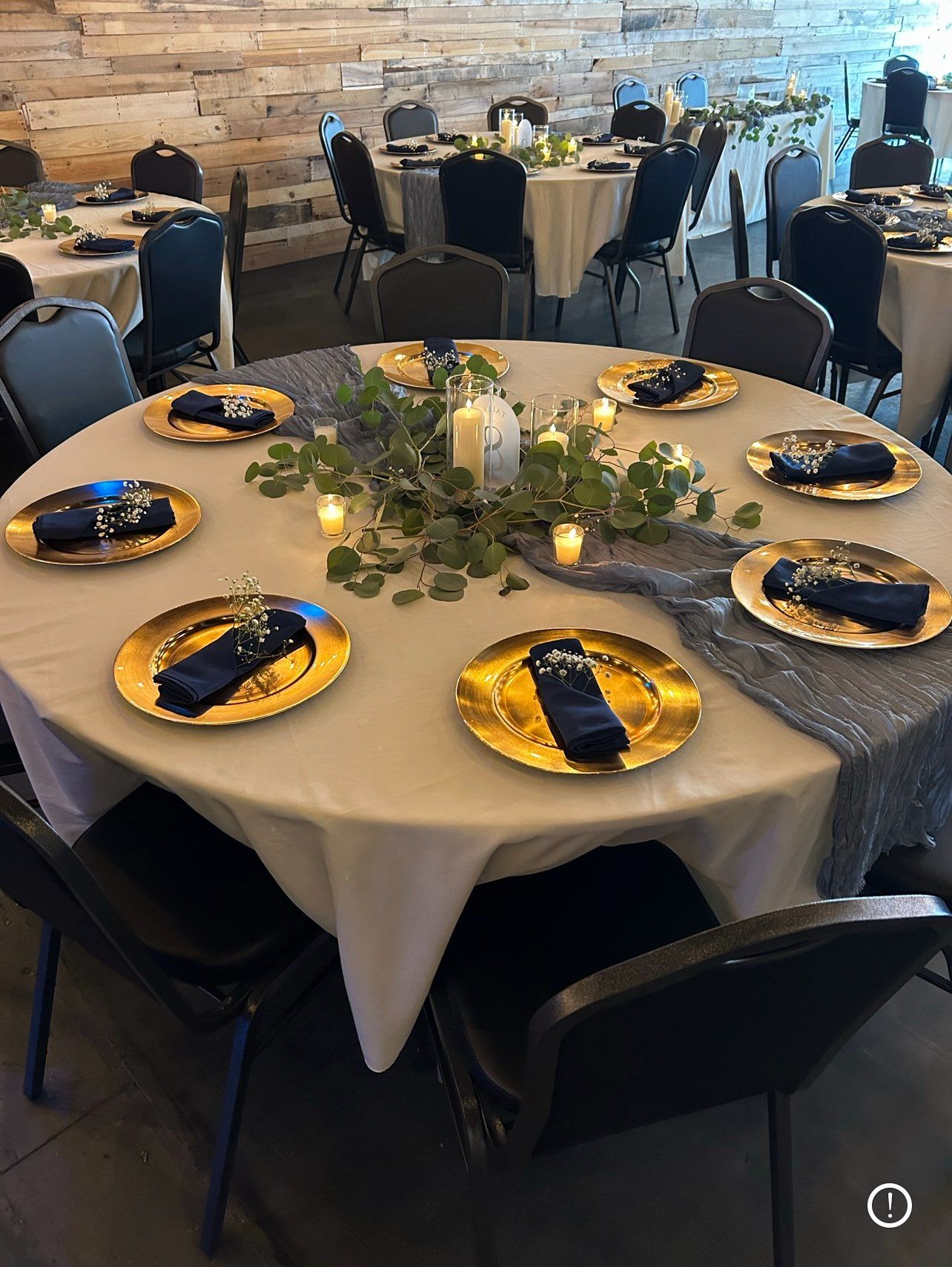 Round table set for a celebration with gold chargers, navy napkins, and greenery centerpiece in a banquet hall.