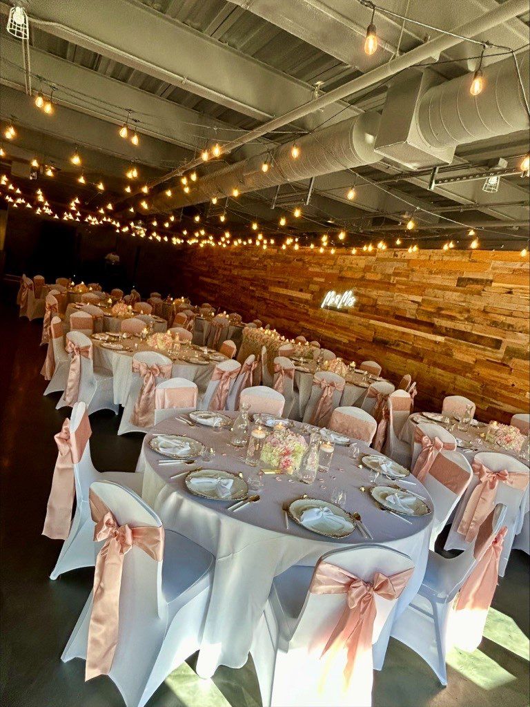 Wedding reception tables with pink bows and centerpieces, under string lights in a brick-walled room.