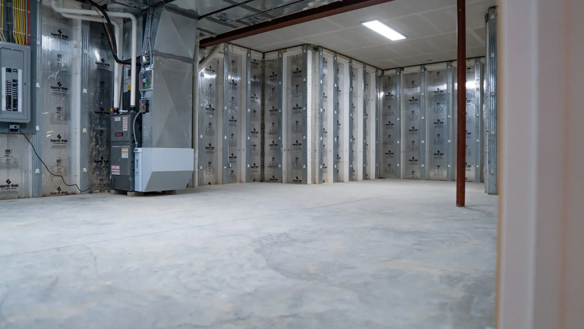 An unfinished basement with concrete floors, metal wall studs with insulation panels, a furnace, and a support pole.