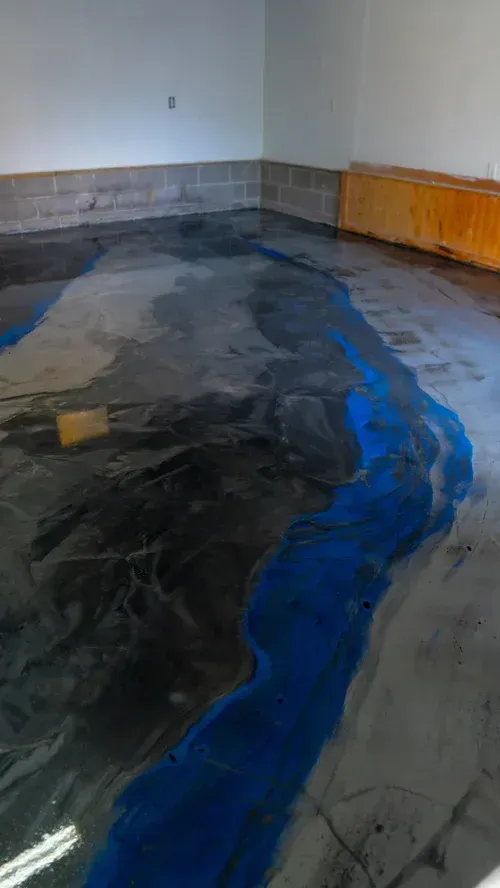 A floor under renovation, with a smooth gray epoxy base featuring a prominent, flowing deep blue river-like inlay.