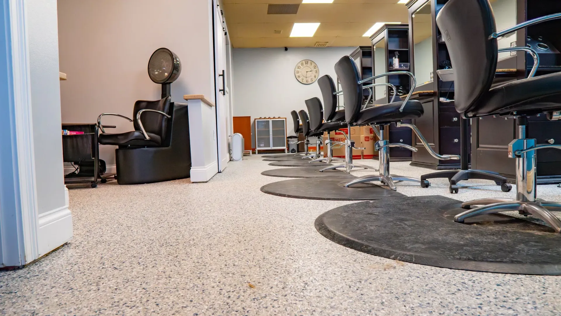 An interior view of a hair salon featuring a row of styling chairs with black floor mats on a speckled light-colored floor.