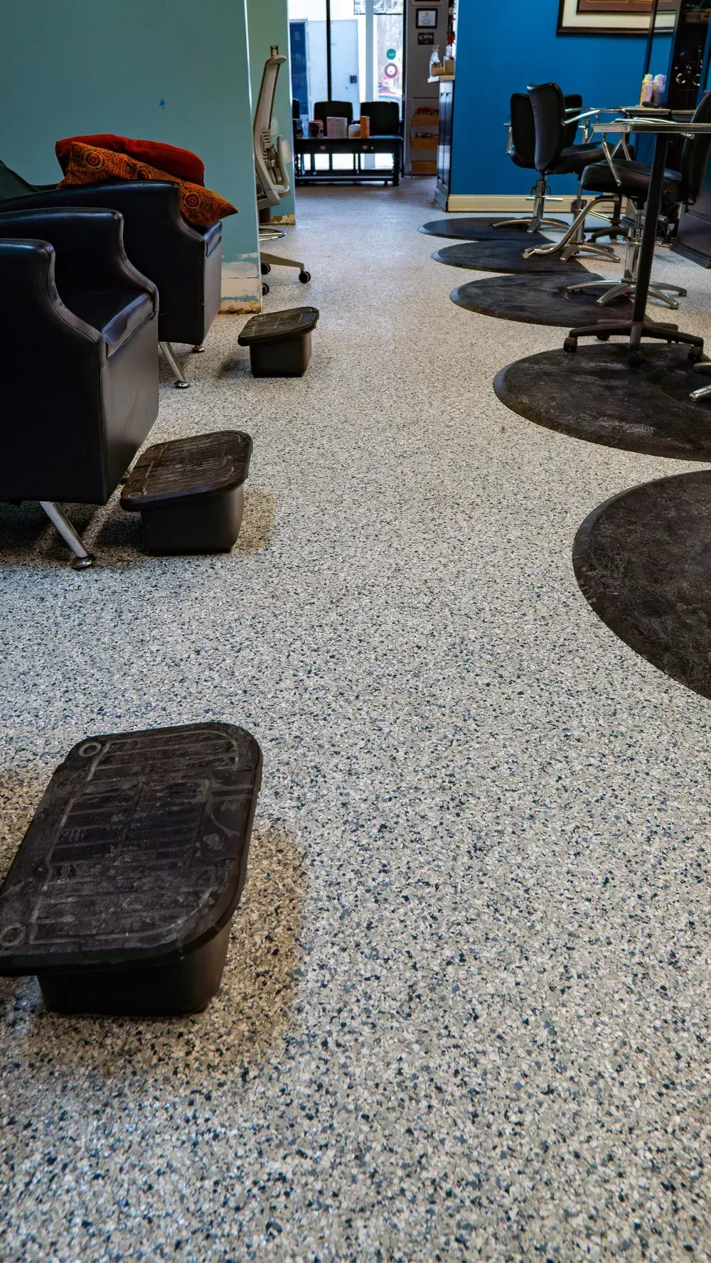 A hair salon interior with a speckled floor, black chairs, and footrests arranged along a narrow aisle.