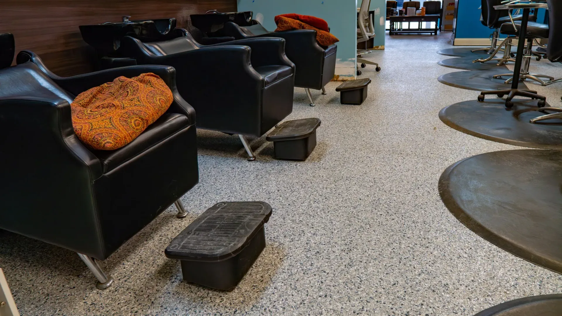 A row of black salon chairs with orange textured cushions on top, arranged for pedicure services in a salon.