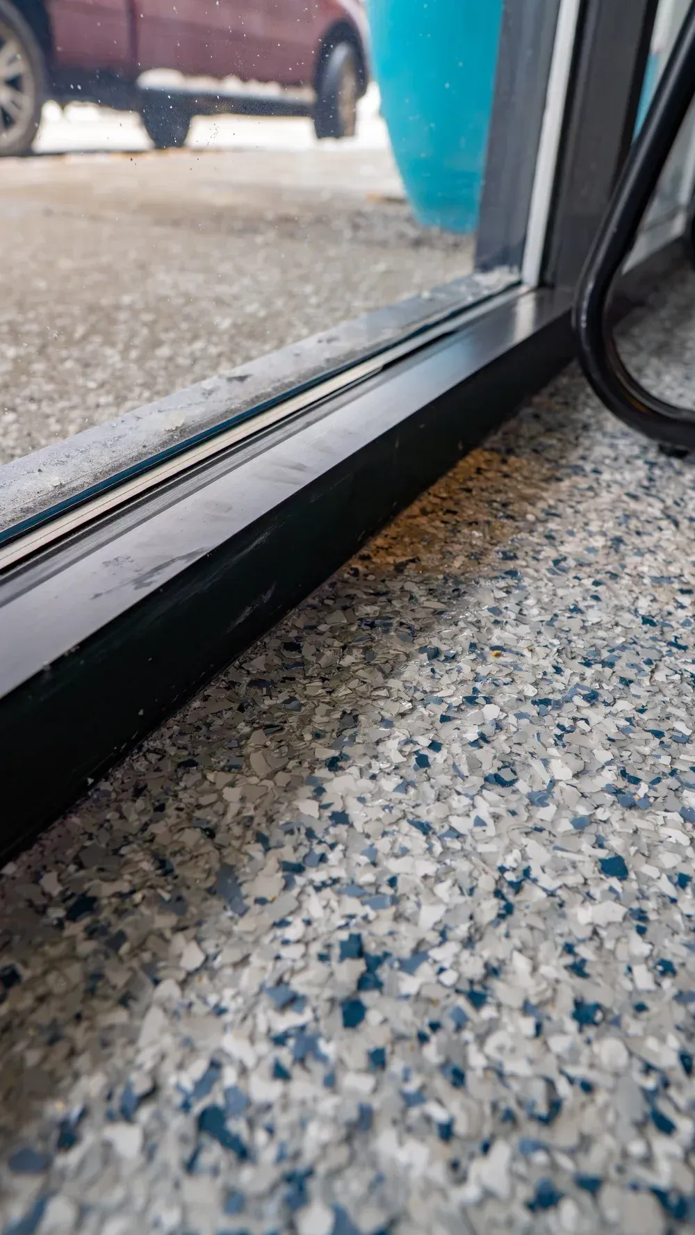 A close-up view of the black metal frame of a glass door sitting above a speckled grey and blue floor.