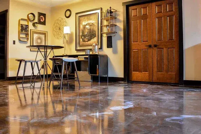 A room featuring a glossy, marbled concrete floor, a small metal table with two stools, wall art, and double wooden doors.