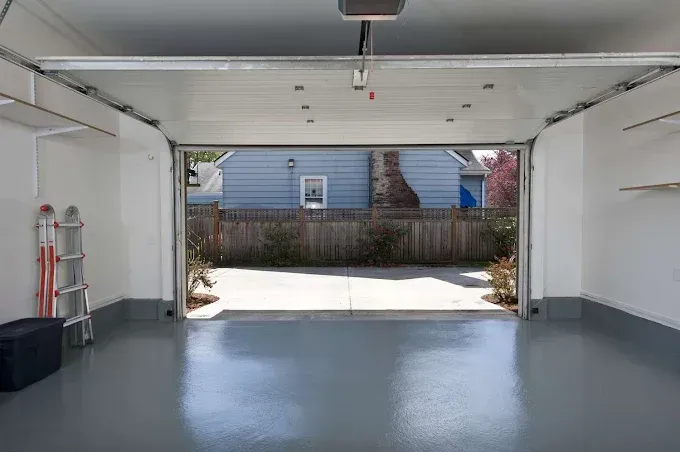 An empty garage with a grey painted floor, white walls, storage shelves, and an open door viewing a backyard.