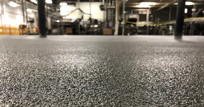A close-up of a gray, textured industrial floor surface in a warehouse or factory setting with out-of-focus equipment.