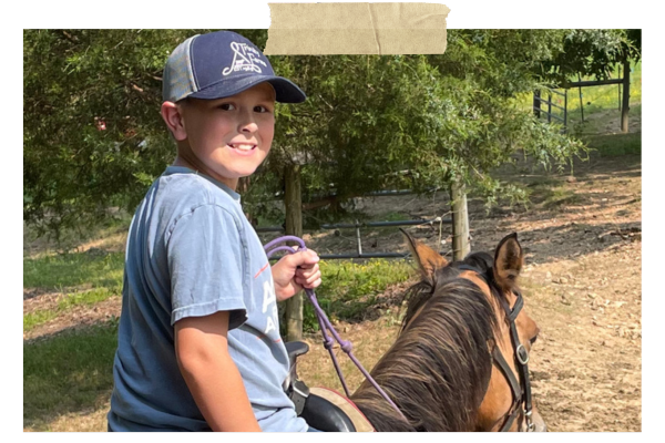 Smiling child on a horse on a wooded trail, wearing a blue shirt and cap.