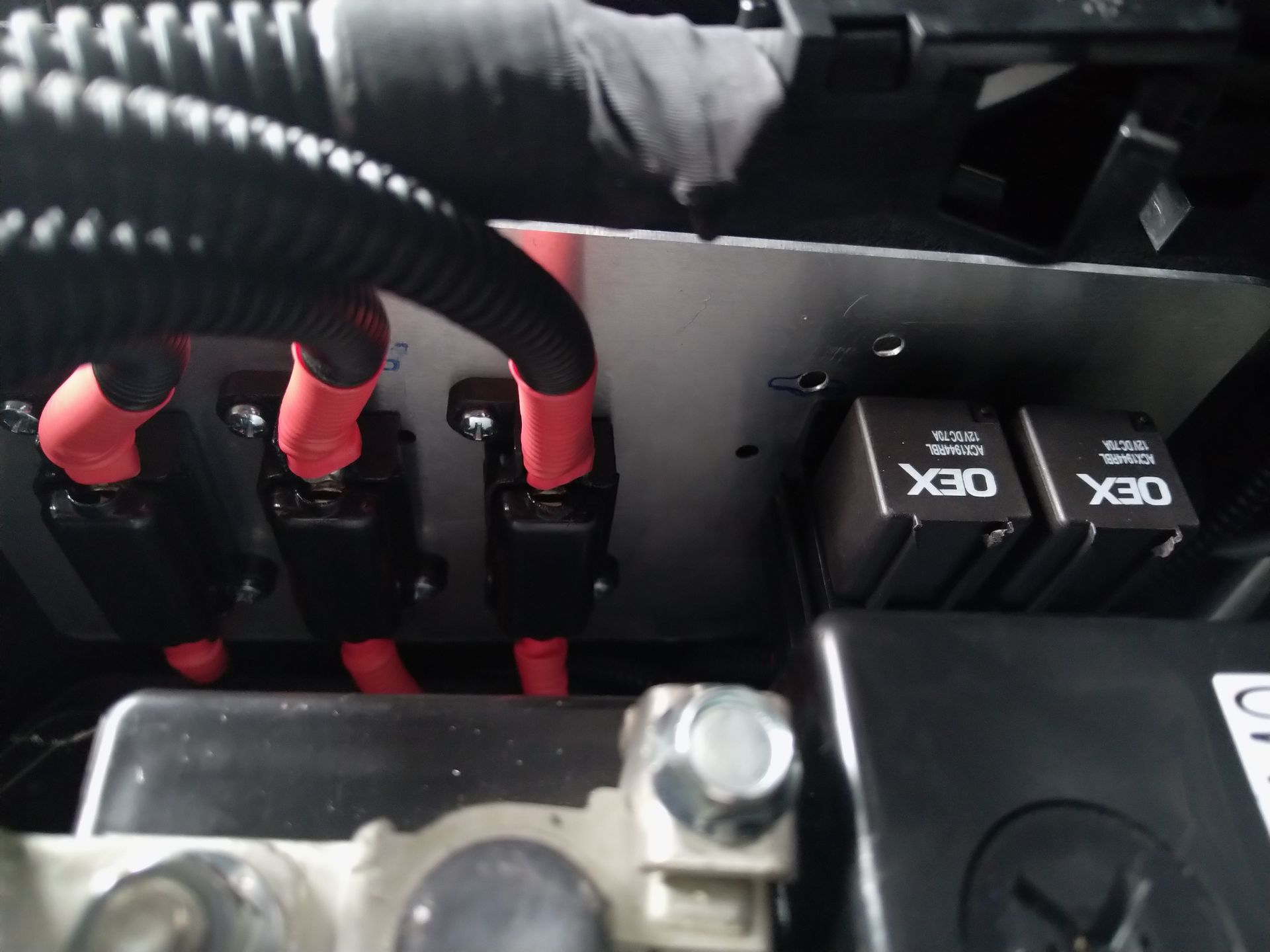 Close-up of car battery terminals and electrical components with red wires, black fuses, and two black relays.