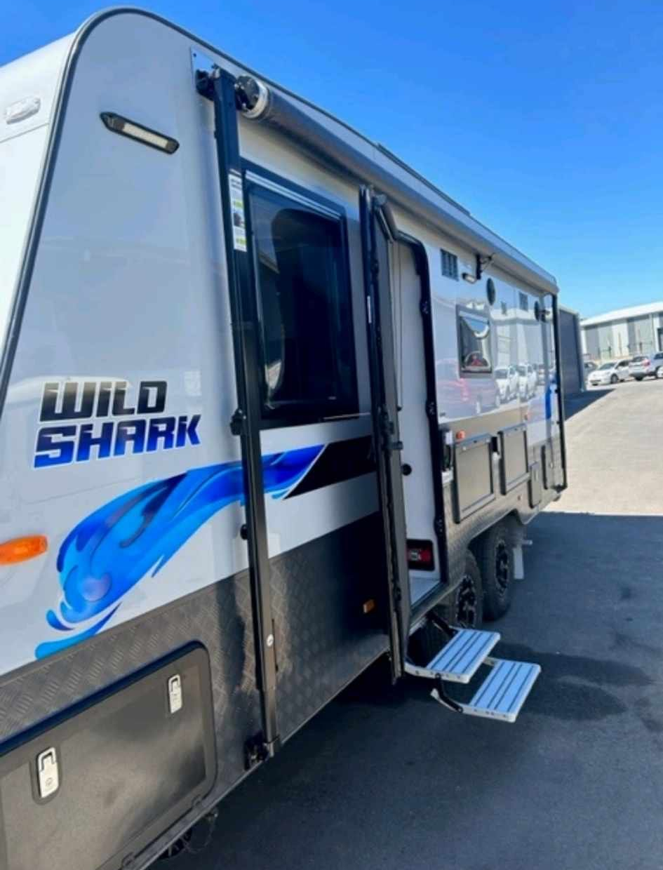 Side view of a Wild Shark caravan, featuring entry steps, door, and awning; parked outside on a sunny day.