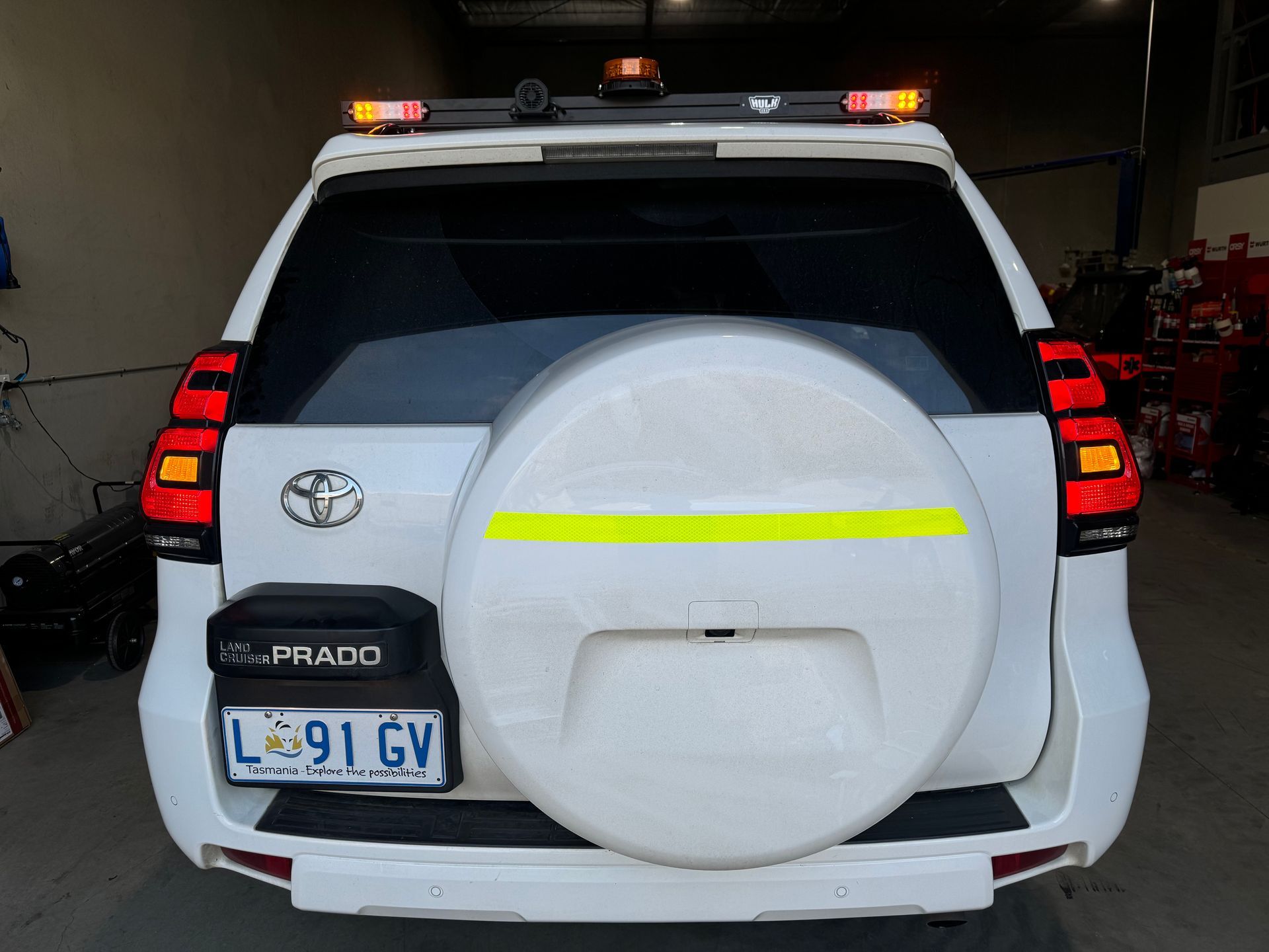 White Toyota Prado SUV with emergency lights on top, spare tire, and reflective tape on the back.