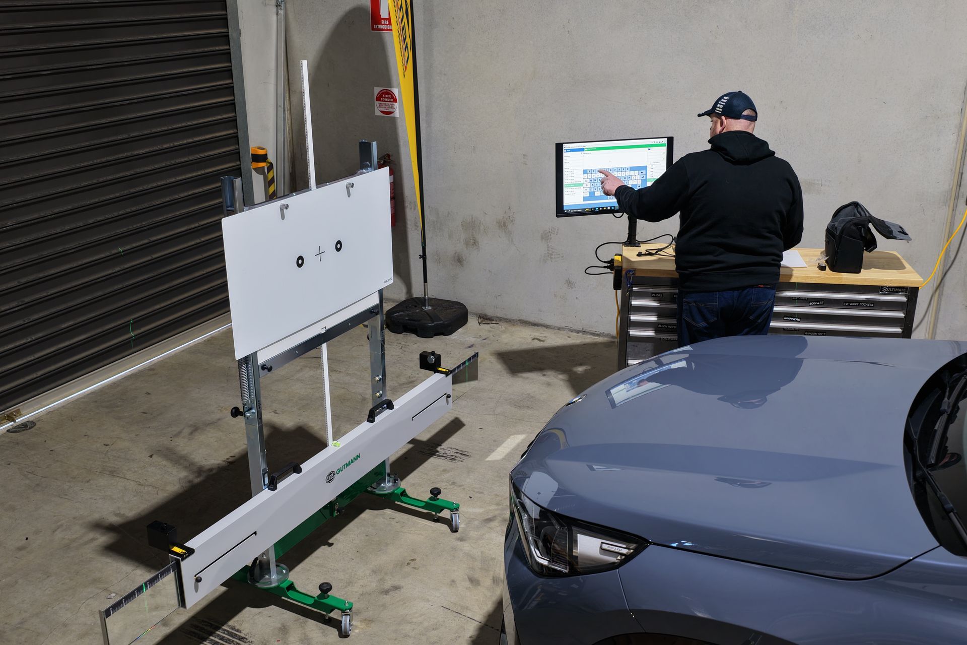 A person calibrating a car's alignment in a garage using a computer and measuring tools.