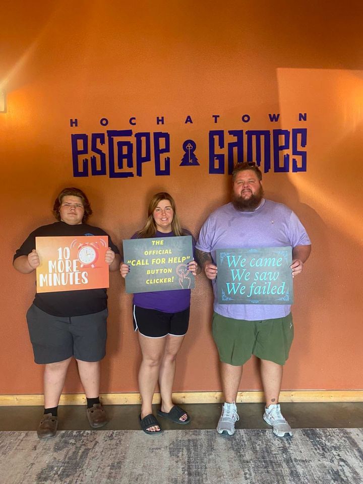 Three people holding colorful signs in front of a “Moktown Escape & Games” wall.
