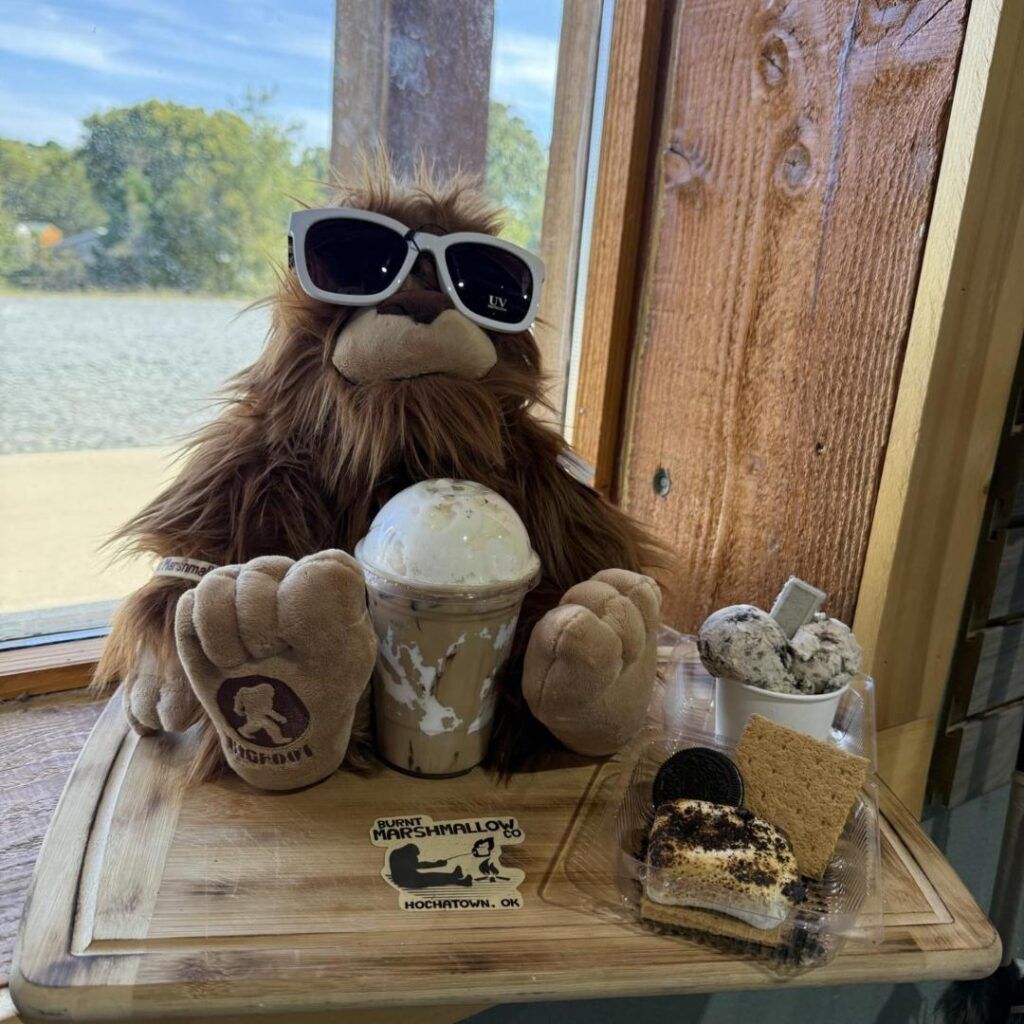 Plush Bigfoot wearing sunglasses beside ice cream treats on a wooden table by a lake view