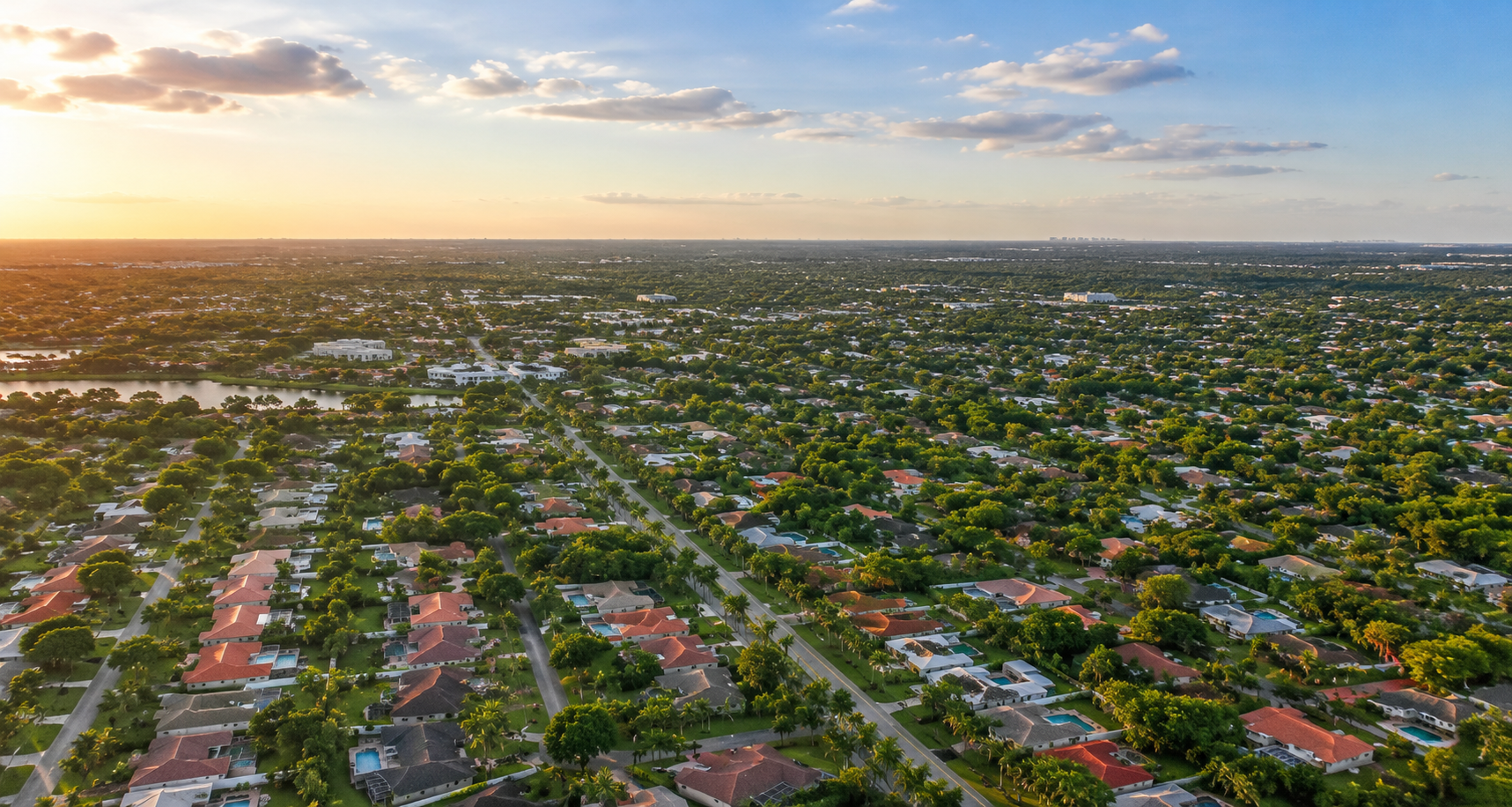 Aerial view of a sprawling suburban neighborhood at sunset with rows of houses and dense green trees