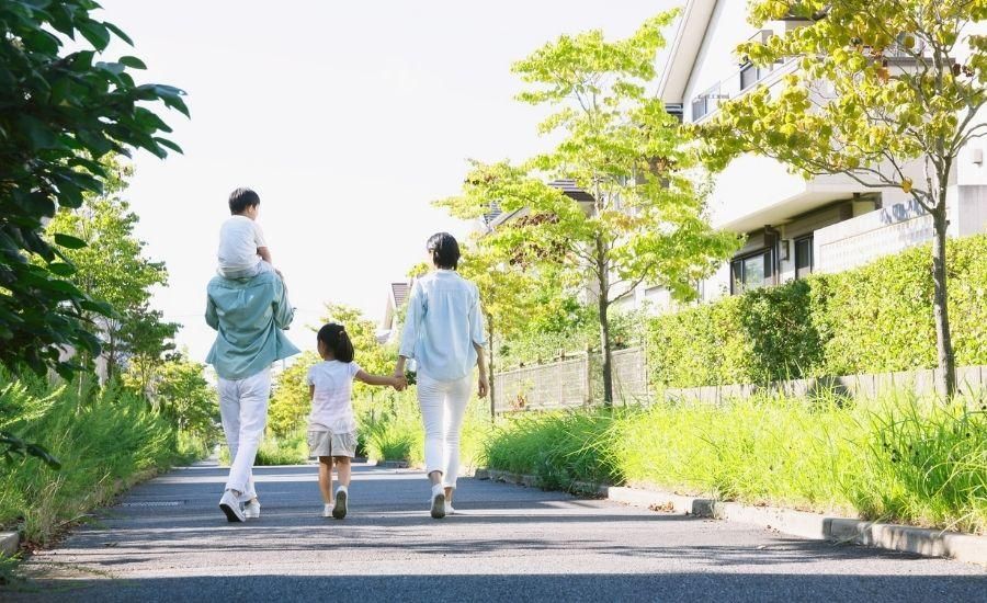 People walking along a tree-lined path on a sunny day, with green hedges and a building nearby