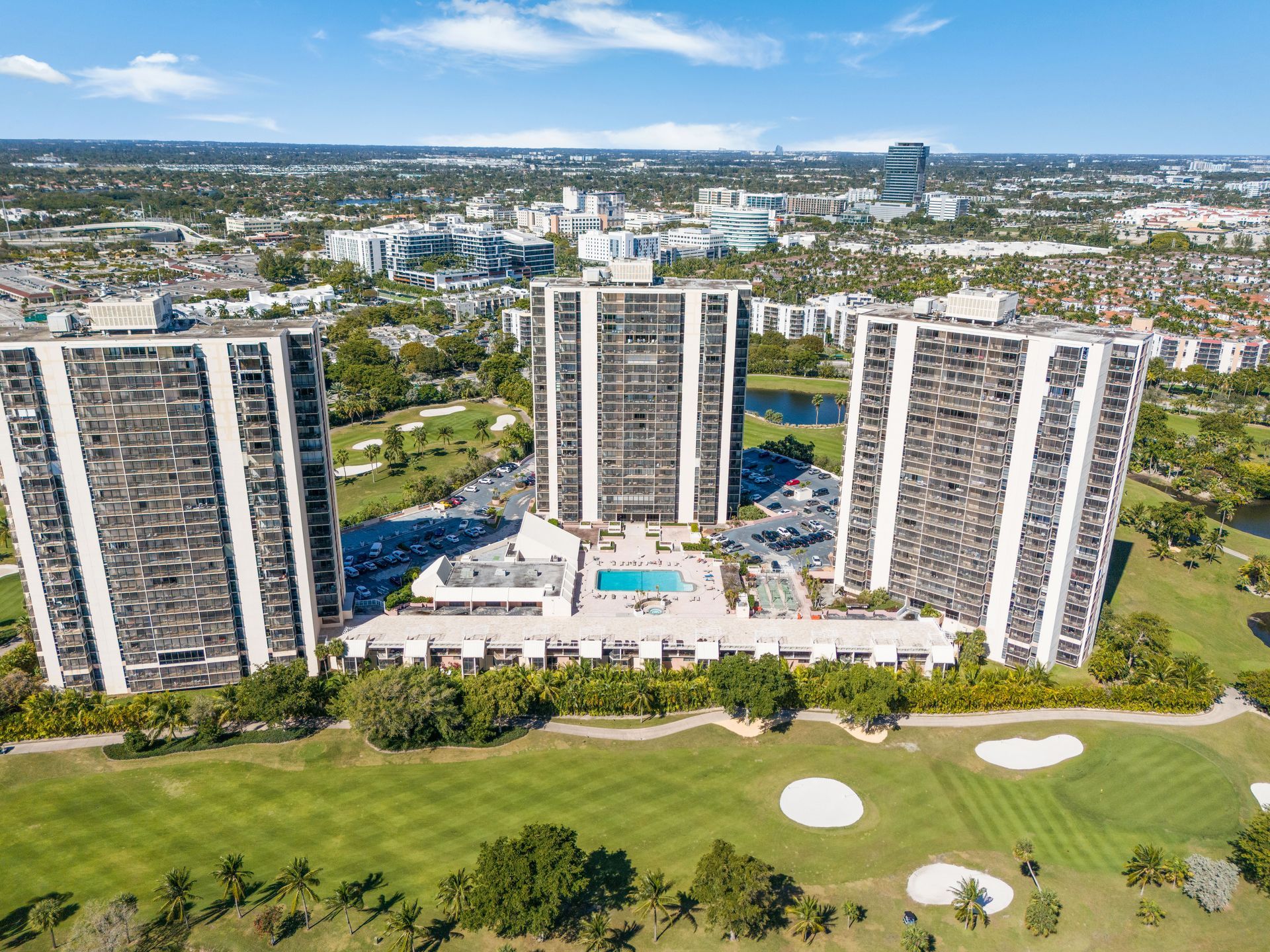 Aerial view of three white high-rise buildings around a central pool and green golf course under blue sky