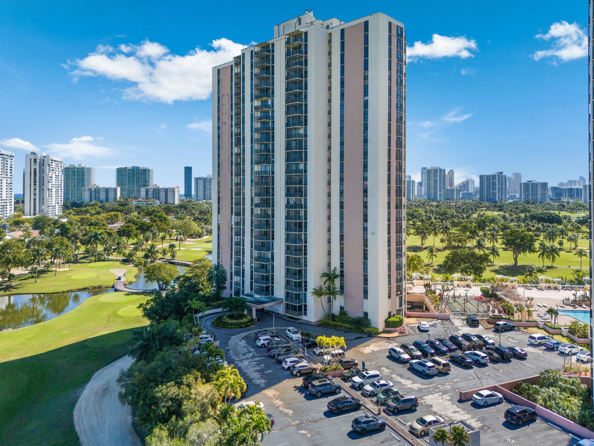 Tall apartment building beside a parking lot and green park under a blue sky with clouds