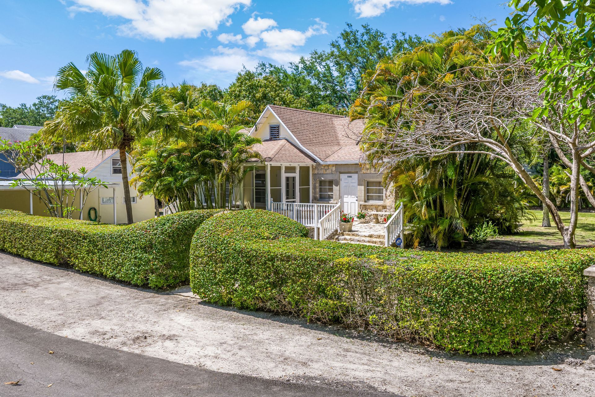 A small white house with a red roof, surrounded by green hedges and palm trees on a sunny day.