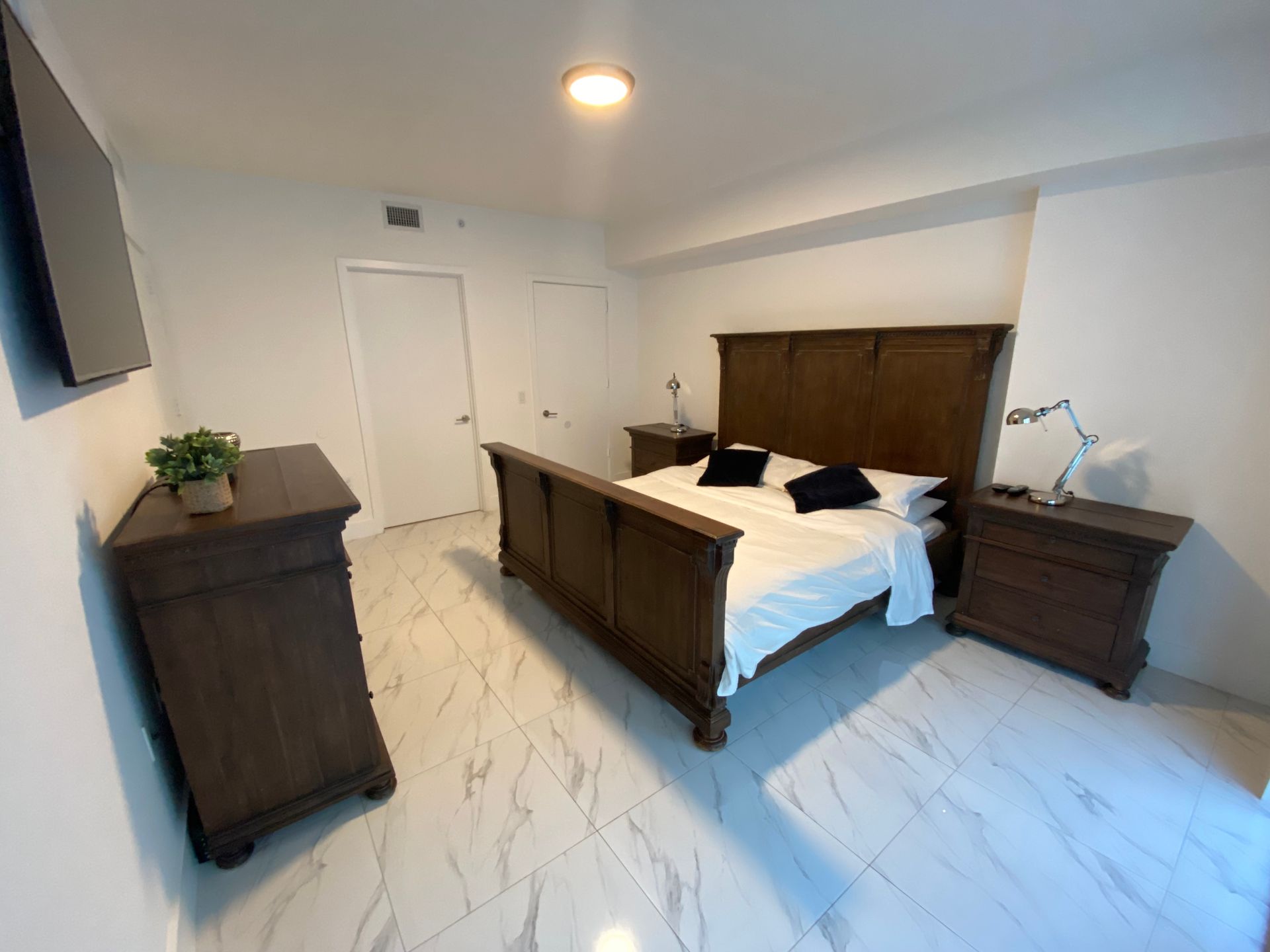 Modern bedroom with dark wood bed, dresser, nightstands, white marble floor, and ceiling fan.