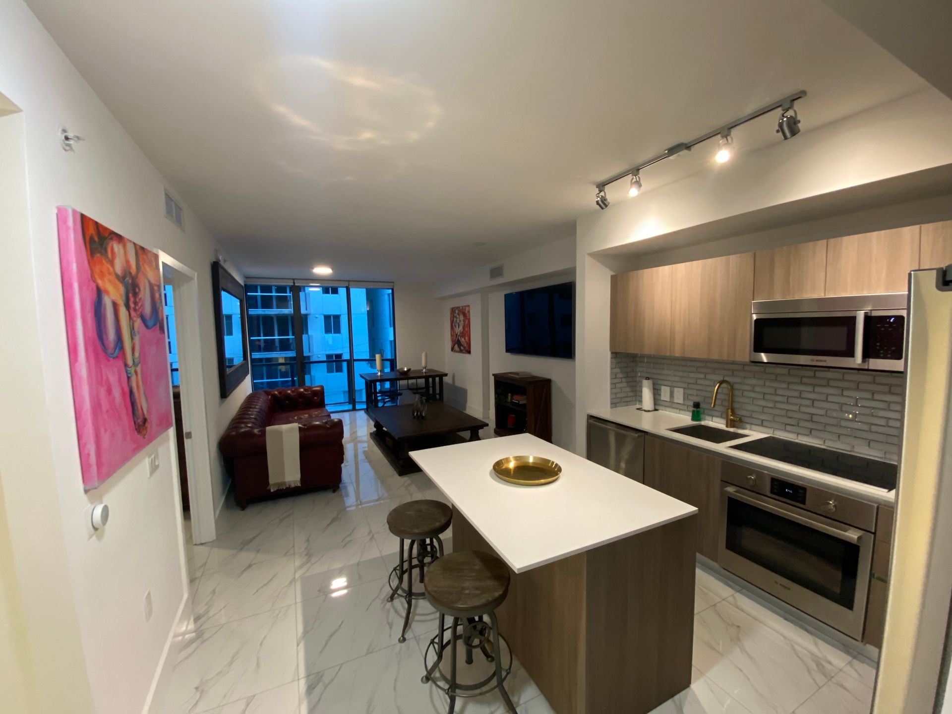 Modern open-plan kitchen and living room with island, stools, stainless appliances, and couch by blue-lit windows
