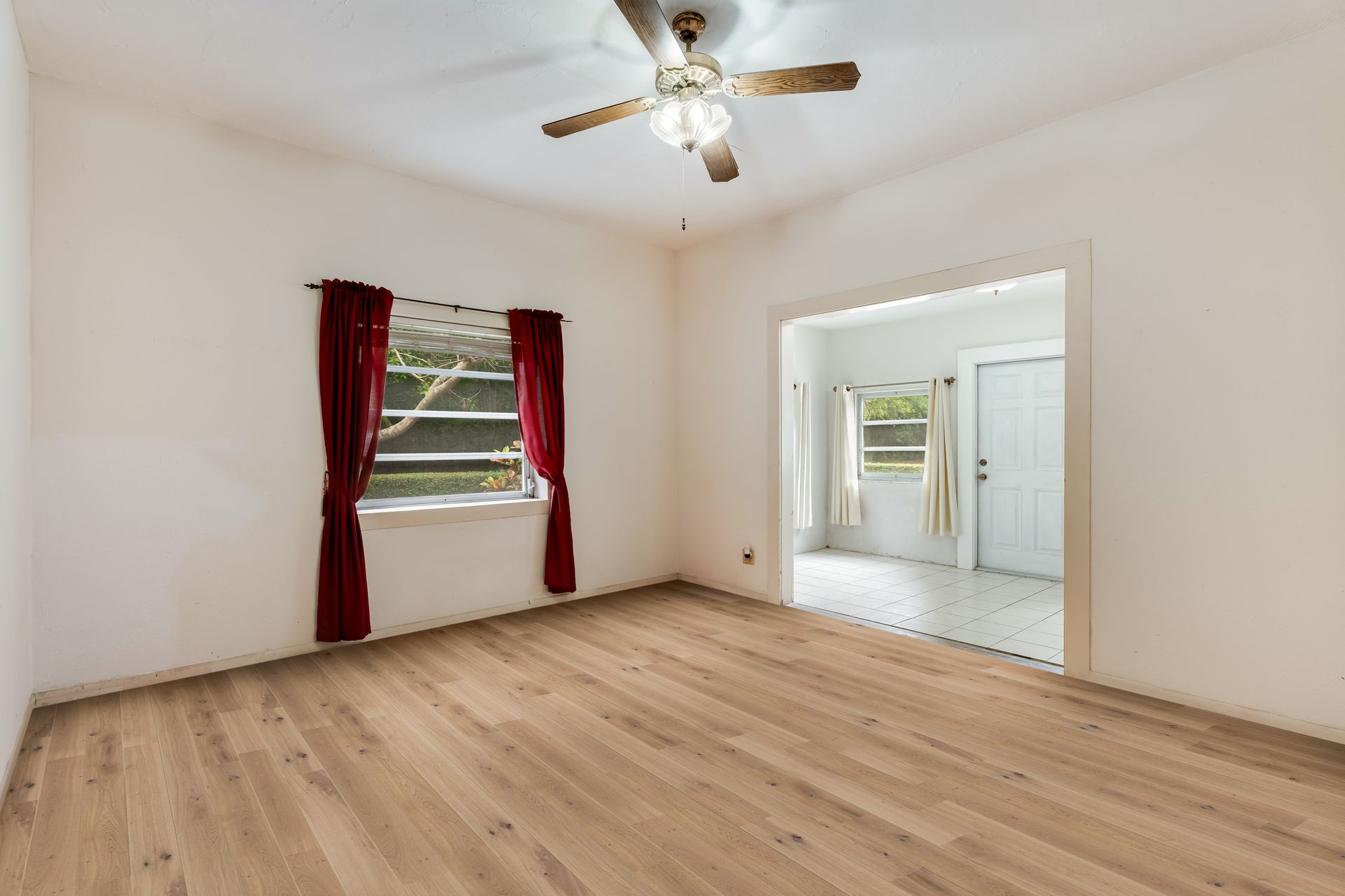 Empty room with wood floors, white walls, ceiling fan, red-curtained window, and open doorway to a bright space