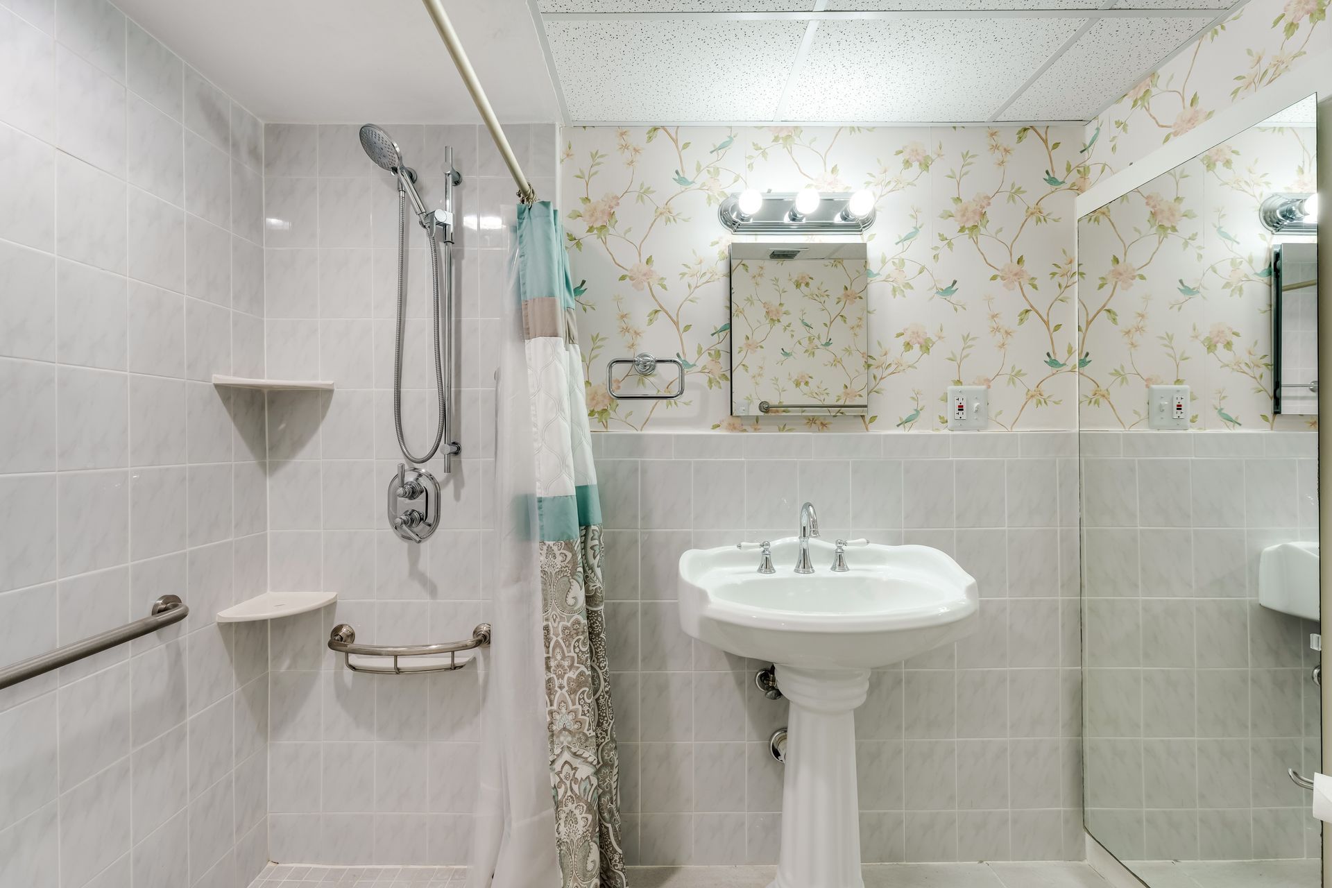 Small bathroom with a shower, pedestal sink, mirror, and white tile walls with floral wallpaper.