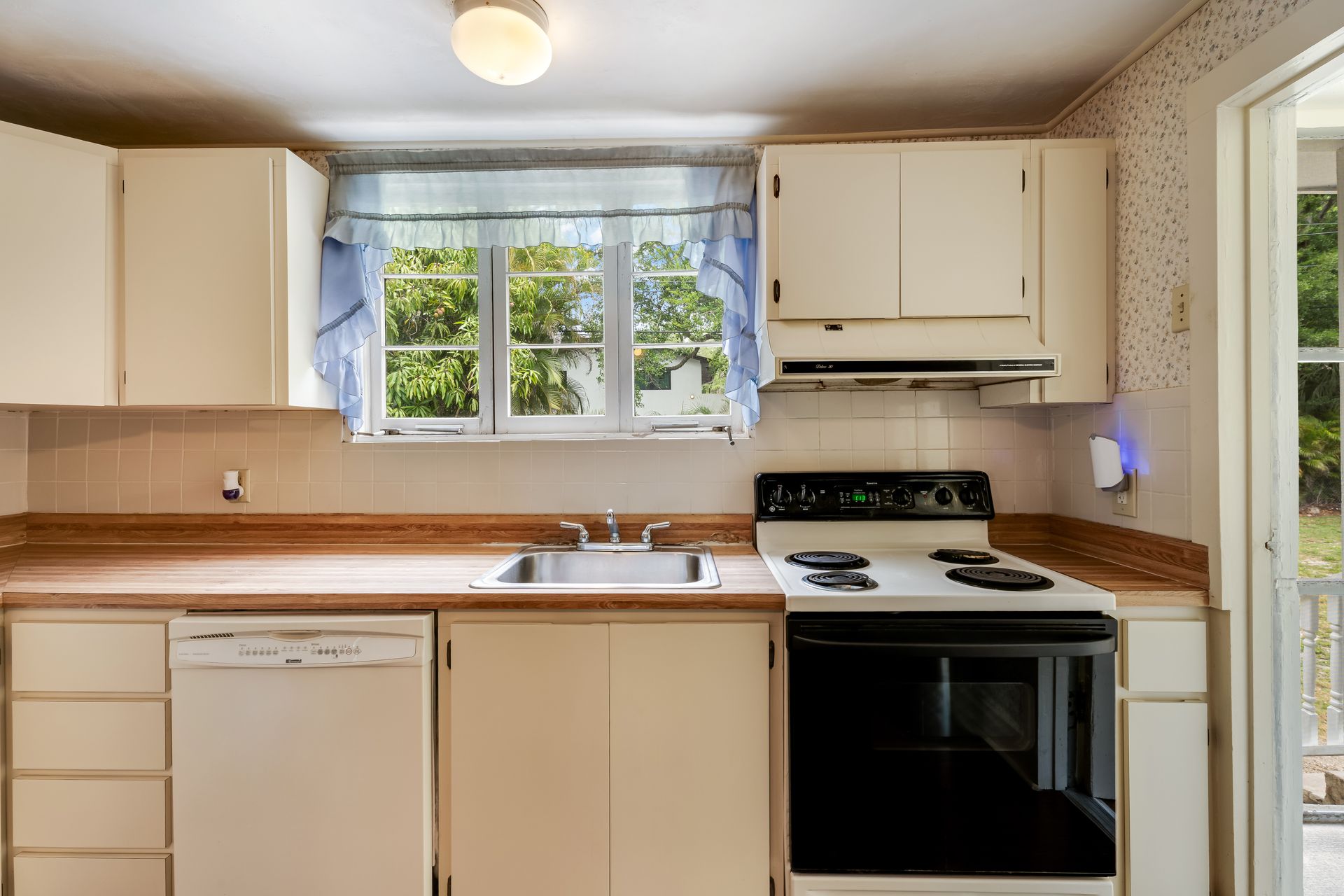 Small beige kitchen with white cabinets, stove, dishwasher, window above sink, and a glass door.