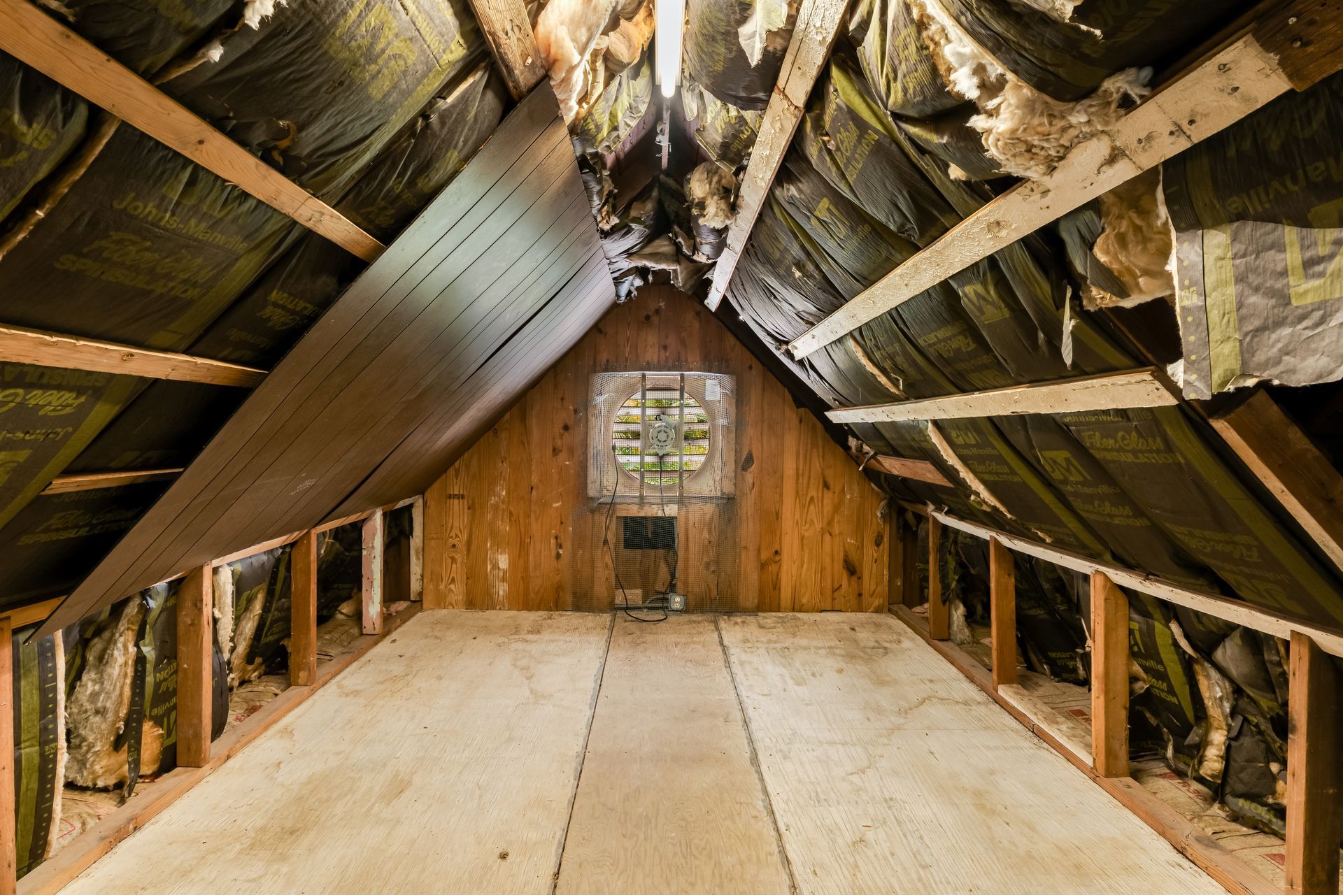 Empty attic with exposed roof beams and a plywood floor, lit by a small window at the far end