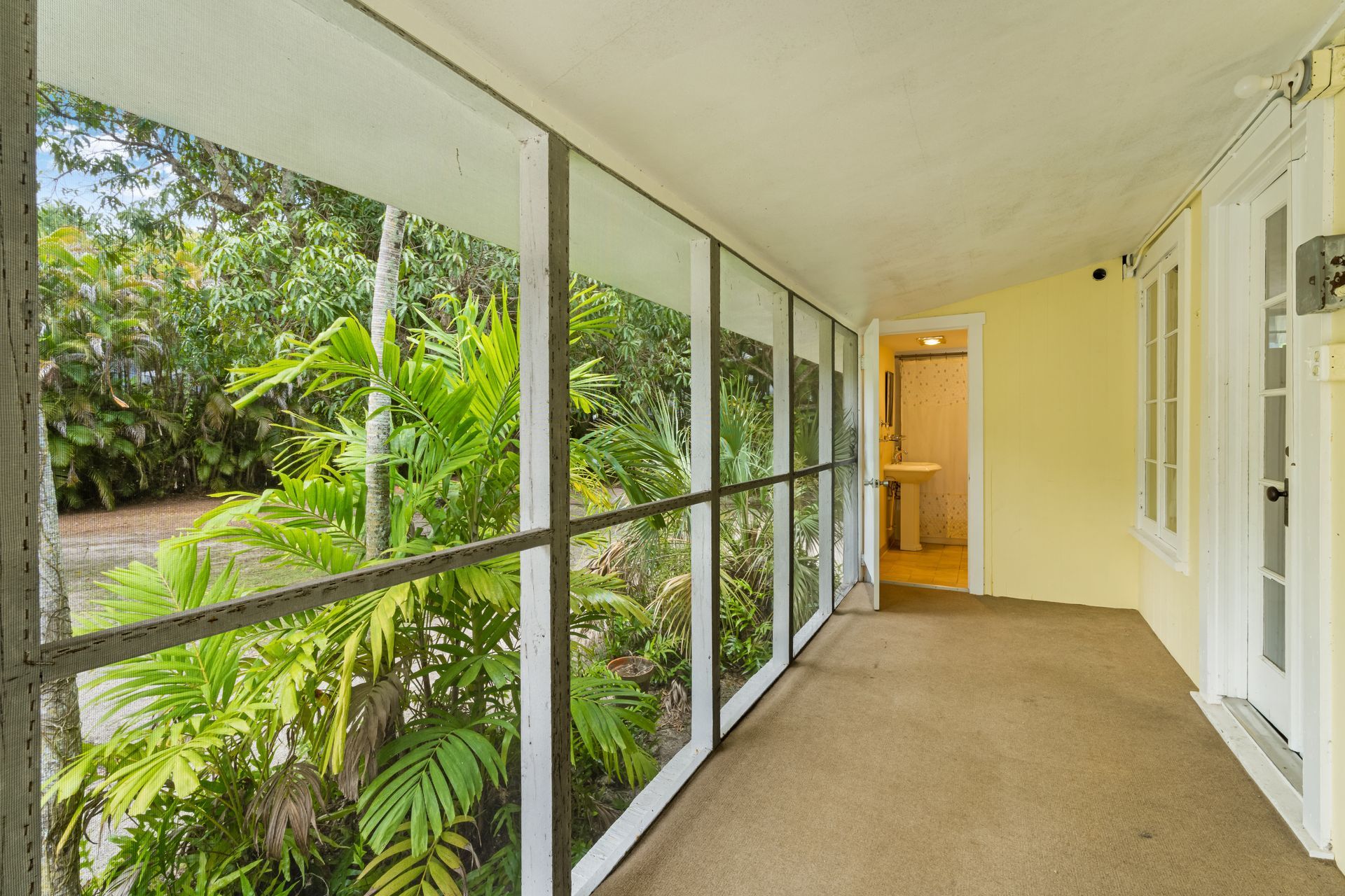 Covered screened porch with carpet, white railing, and a doorway overlooking lush greenery