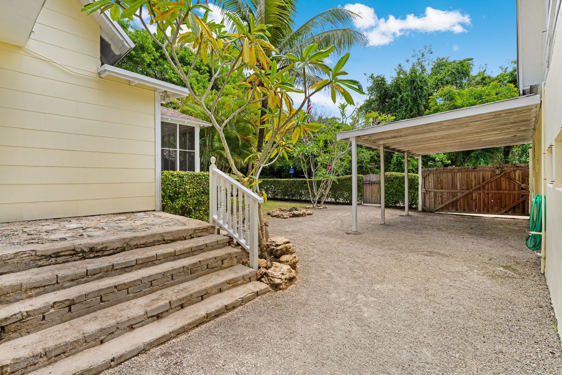Gravel backyard with steps to a cream house and a covered carport under palm trees