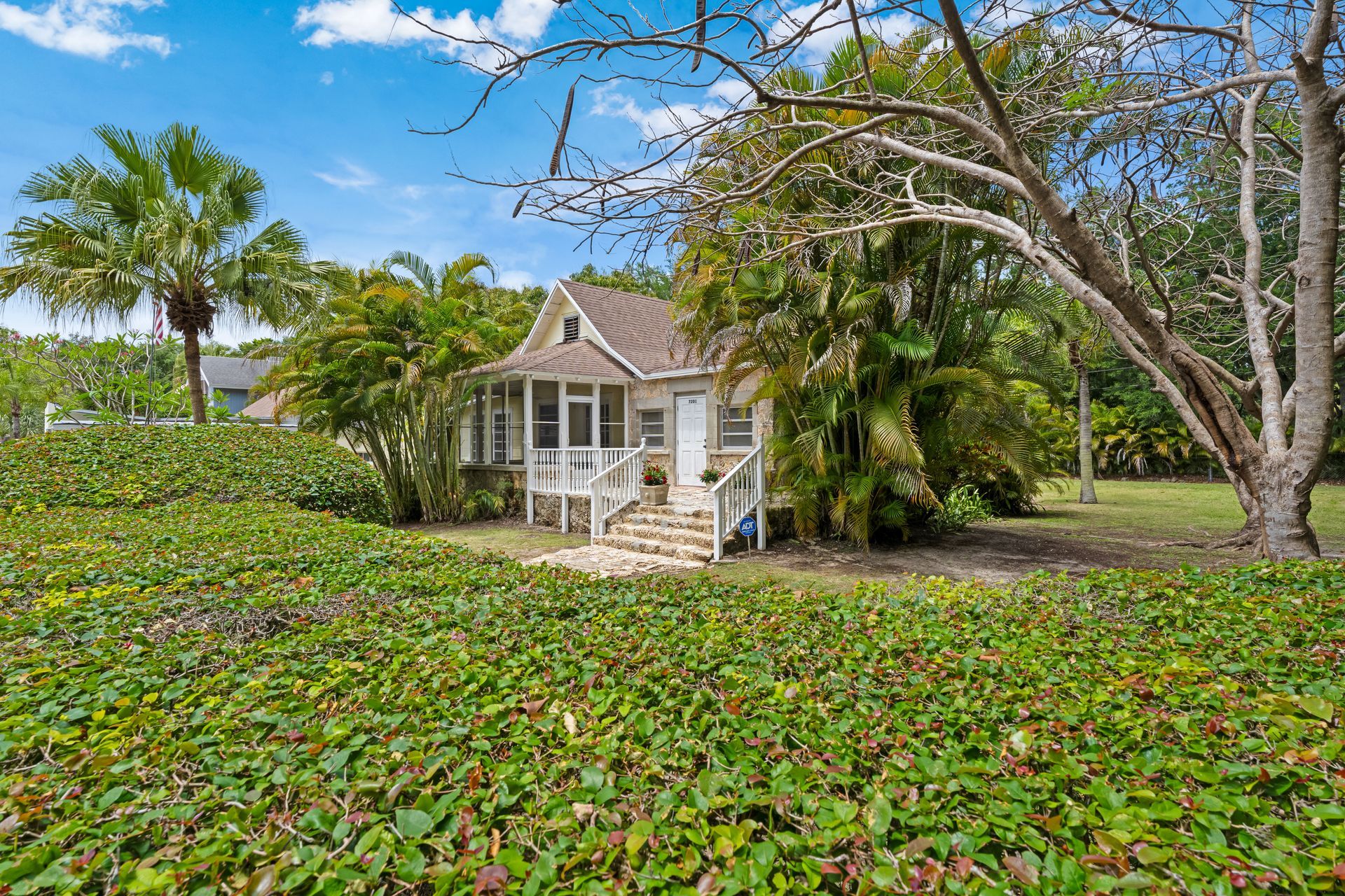 Small white cottage with porch surrounded by lush tropical greenery and palm trees under a blue sky