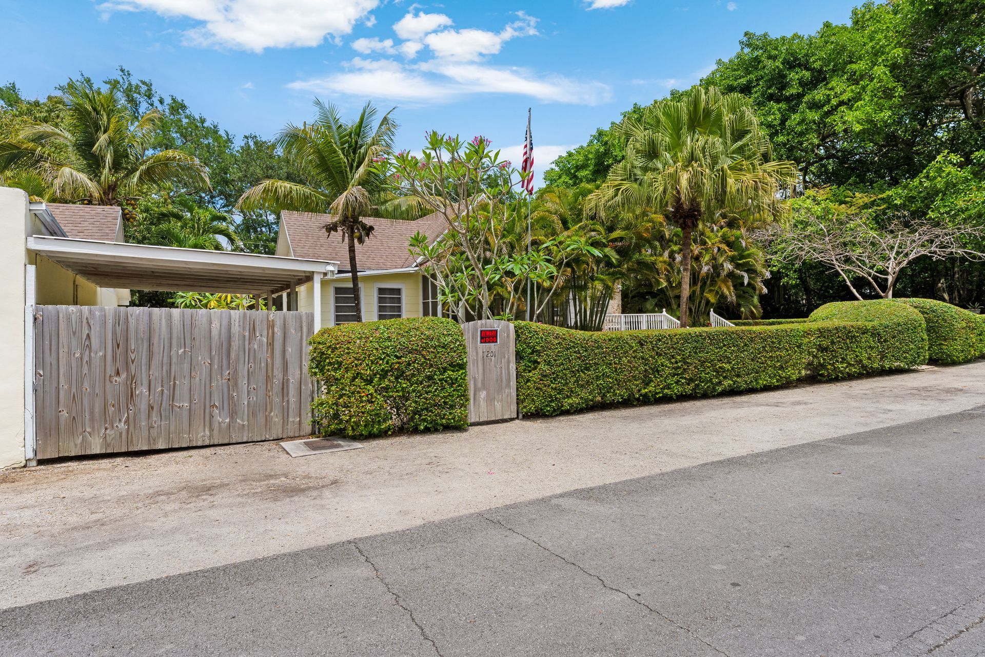 Suburban house behind a wooden gate and hedge, with palm trees and a quiet street in front