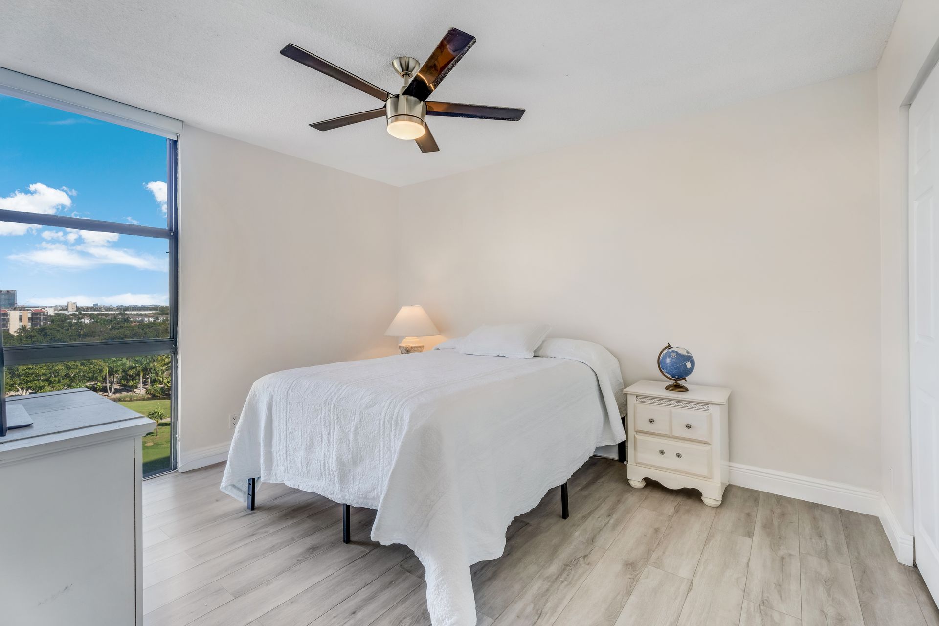 Bright bedroom with a white bed, nightstand, ceiling fan, and ocean view through a large window.