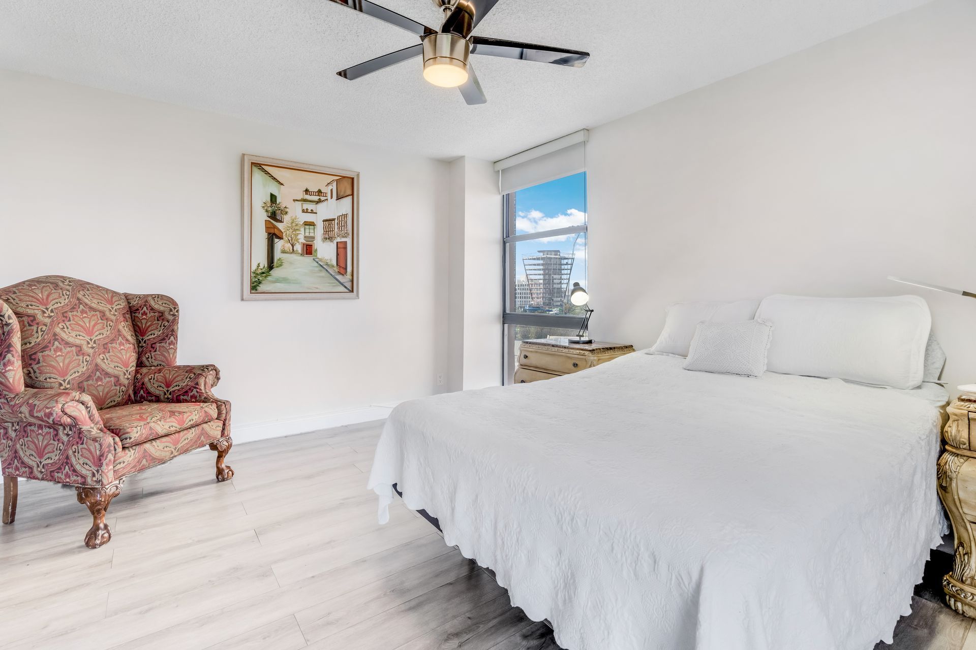 Bright bedroom with white bed, floral armchair, ceiling fan, and window view