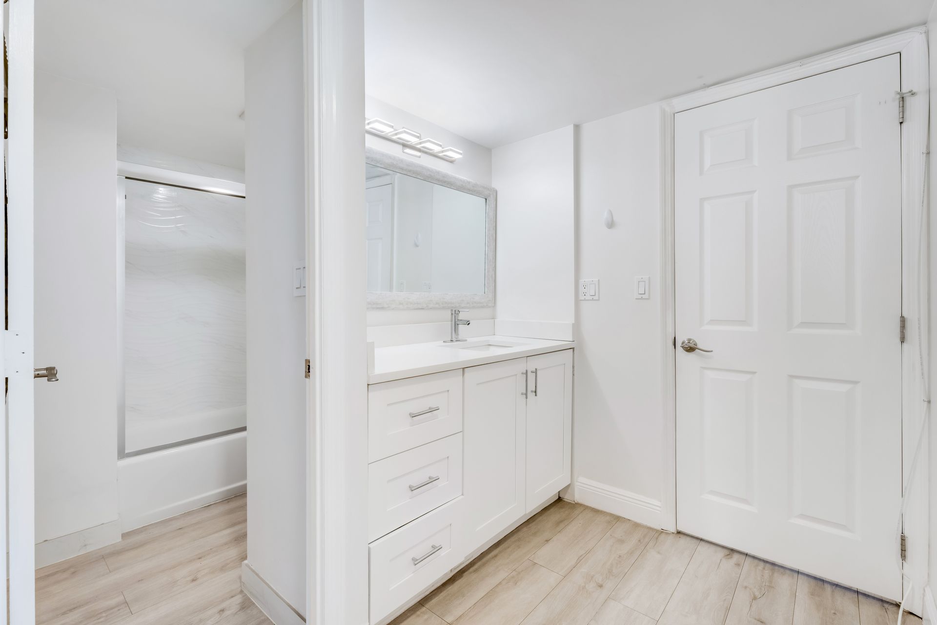 Bright white bathroom vanity with mirror, shower stall, and closed door on light wood flooring.