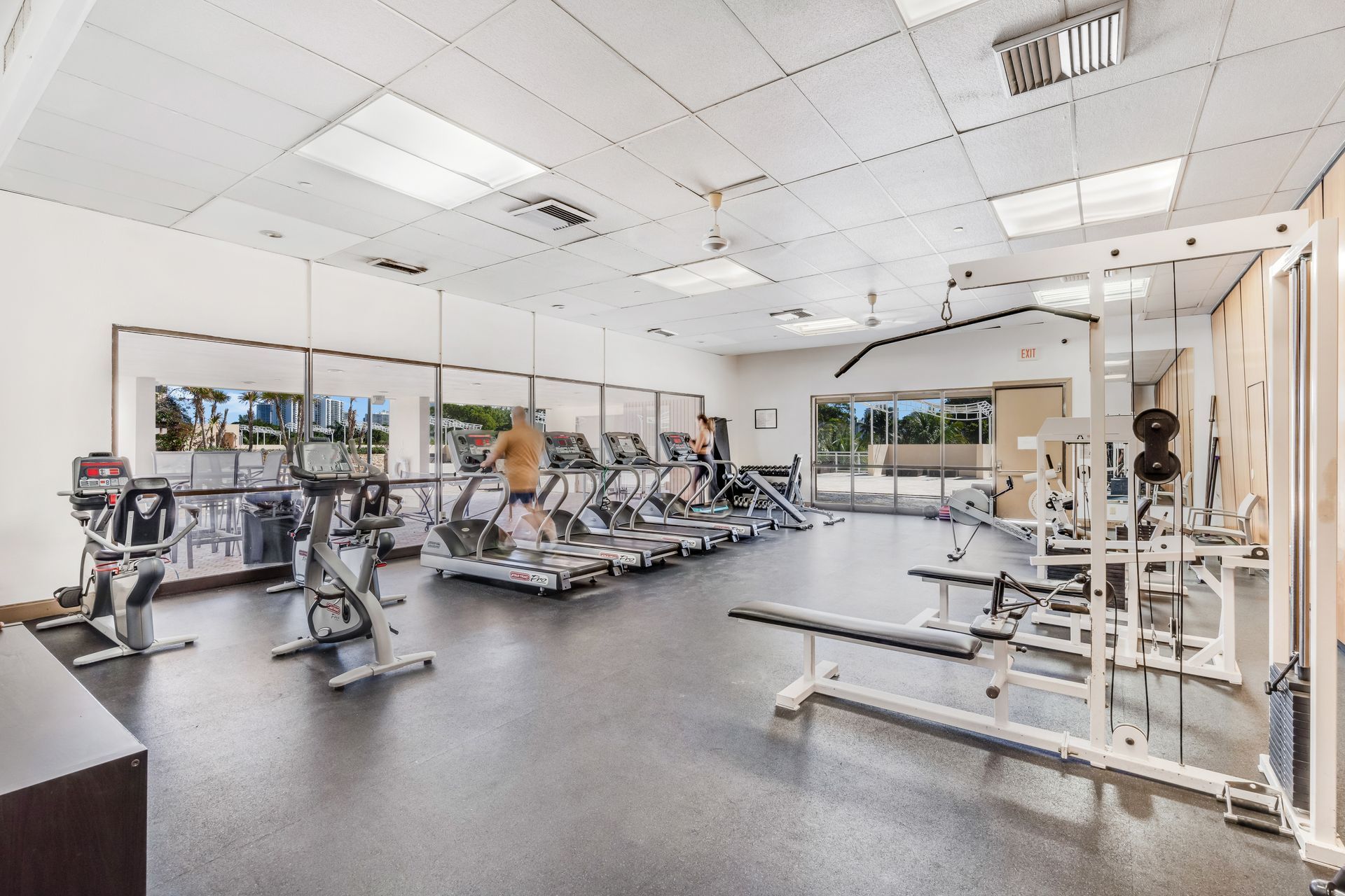 Bright gym interior with rows of exercise machines, weight benches, and mirrors under white ceiling lights