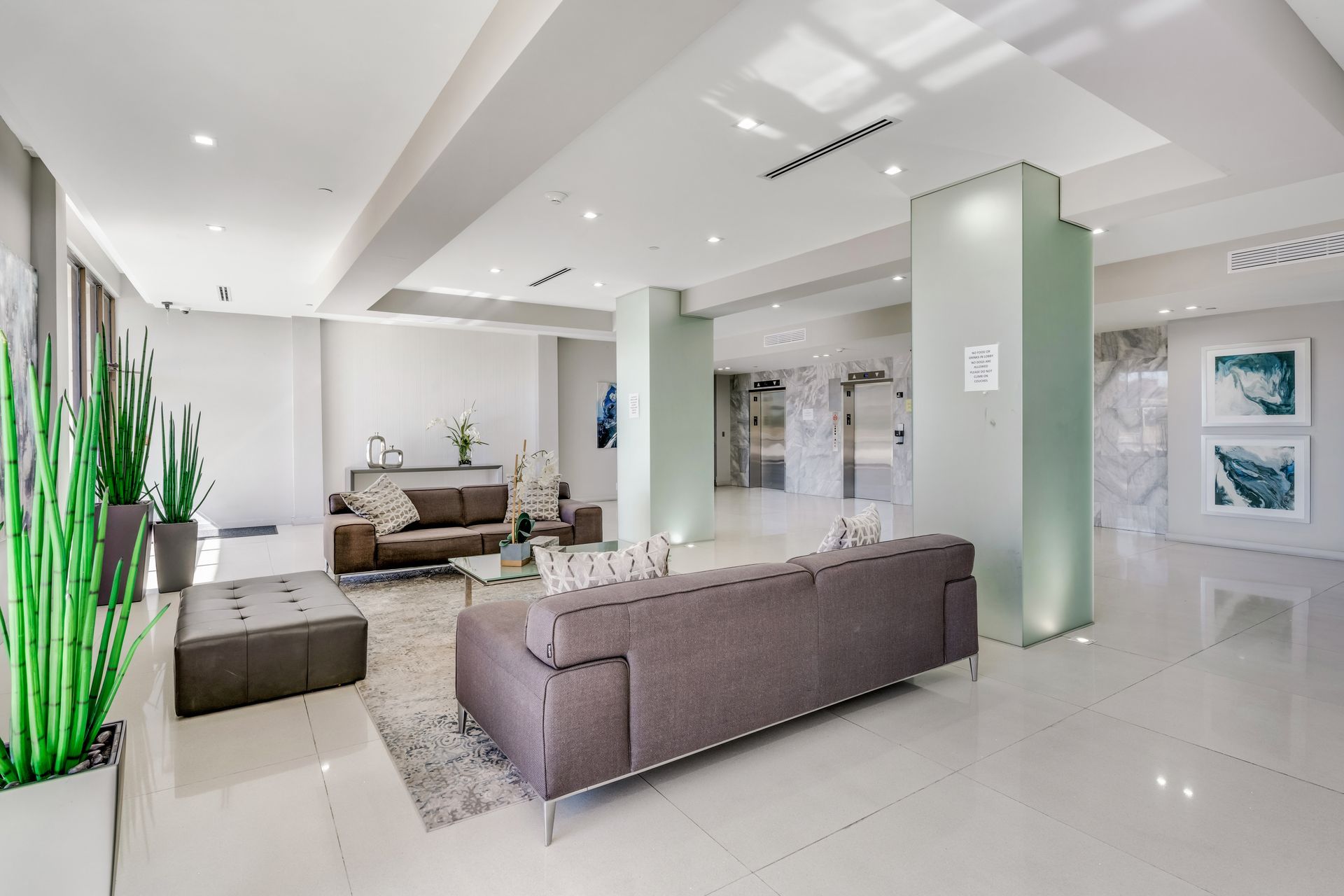 Bright modern lobby with gray sofas, white tile floors, glass columns, and potted plants